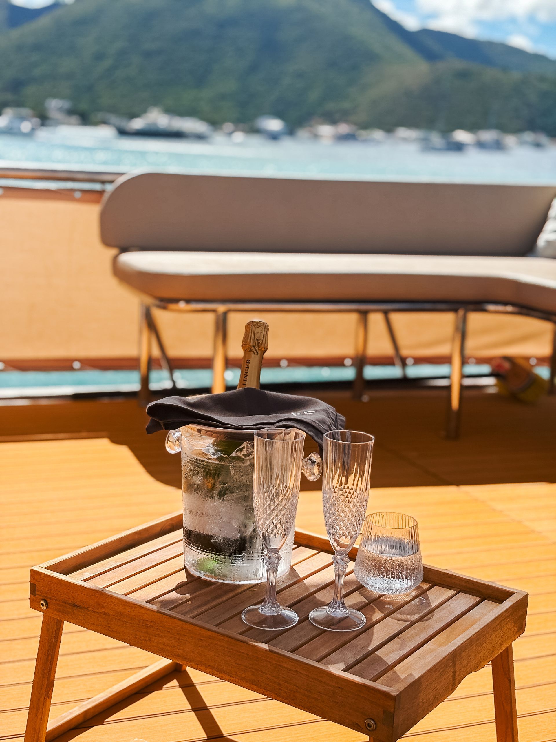 Champagne and glasses on a wooden tray set on a yacht deck, with ocean and mountains in the background.