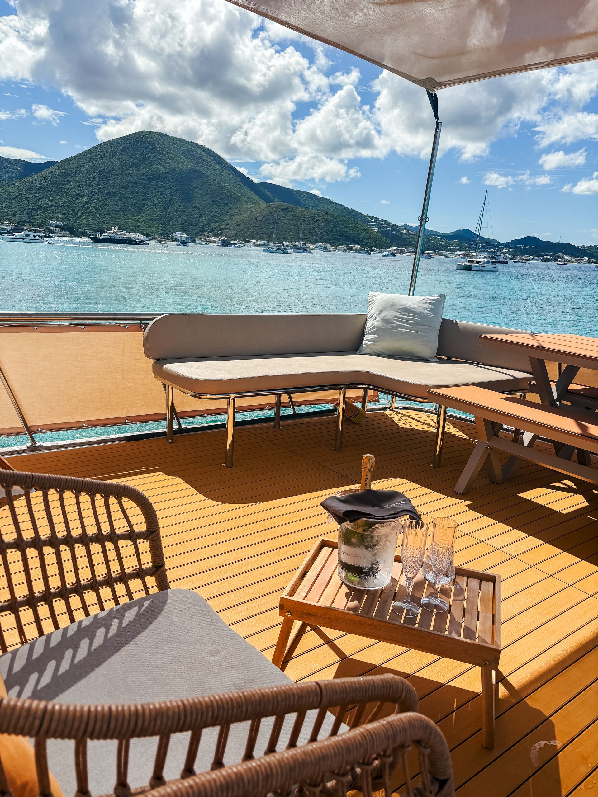 Deck of yacht with seating, bottle in ice bucket, ocean, mountain backdrop.