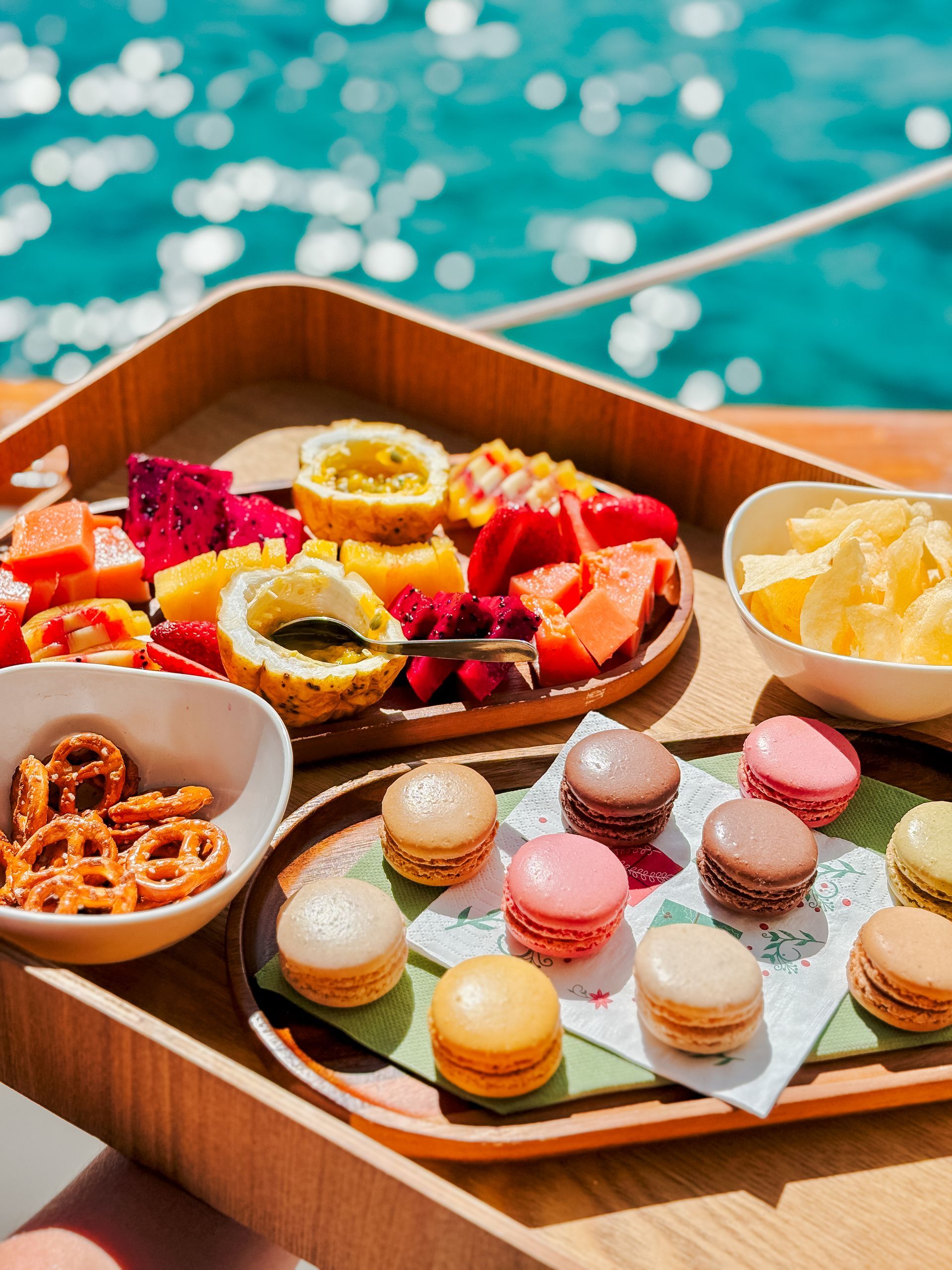 Wooden tray with snacks, including fruit, pretzels, and macarons, on a boat with ocean in the background.