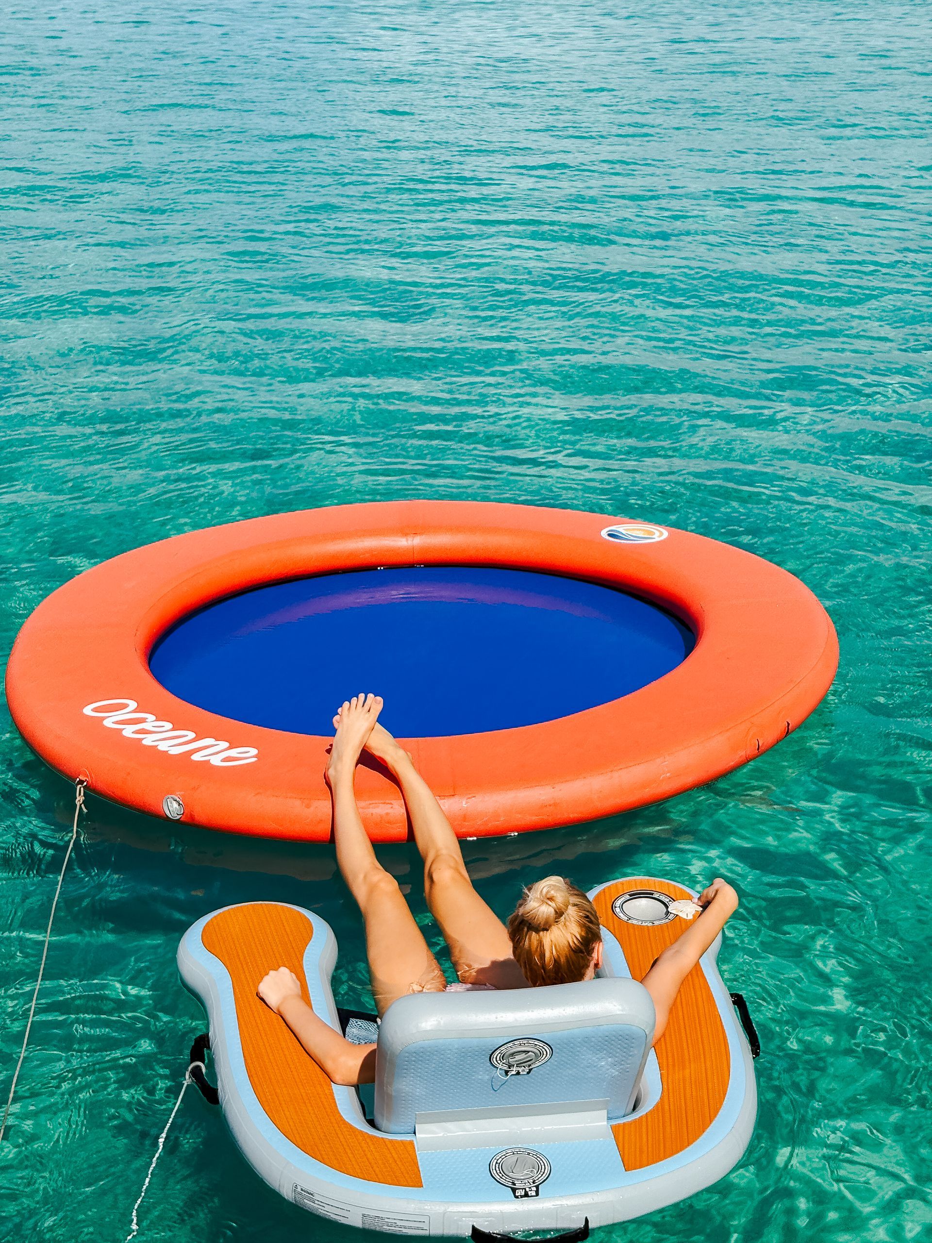 Woman relaxing on a floating lounge chair next to an orange and blue inflatable pool in clear turquoise water.