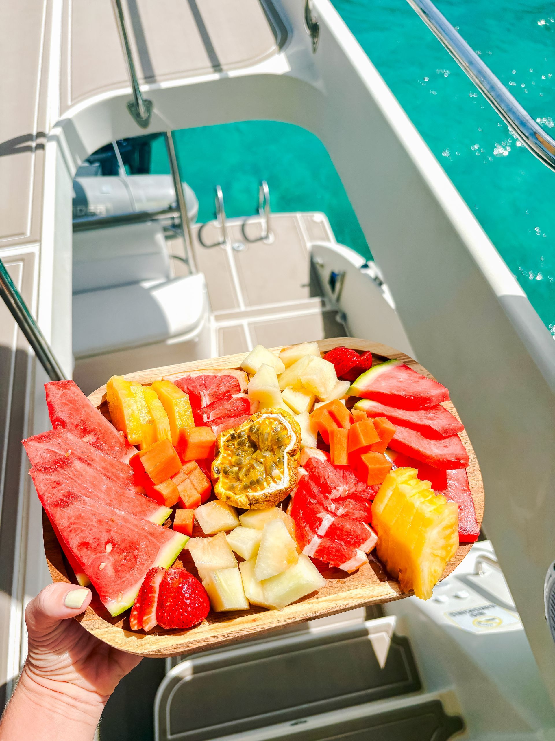 Person holding a fruit platter with watermelon, papaya, and other fruits on a boat with turquoise water.