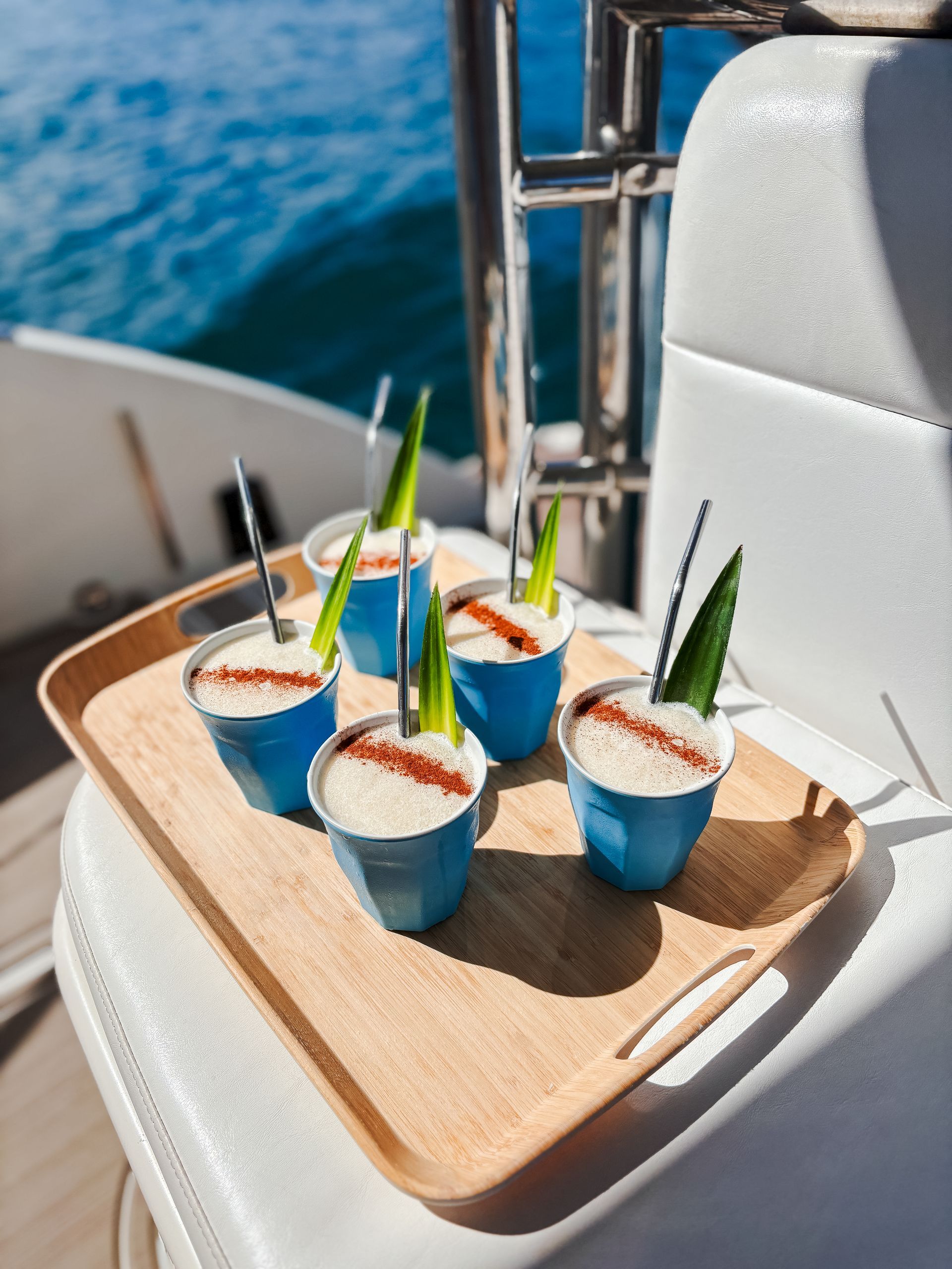 Cocktails in blue cups on a wooden tray, decorated with green sprigs and straws on a boat, ocean in background.
