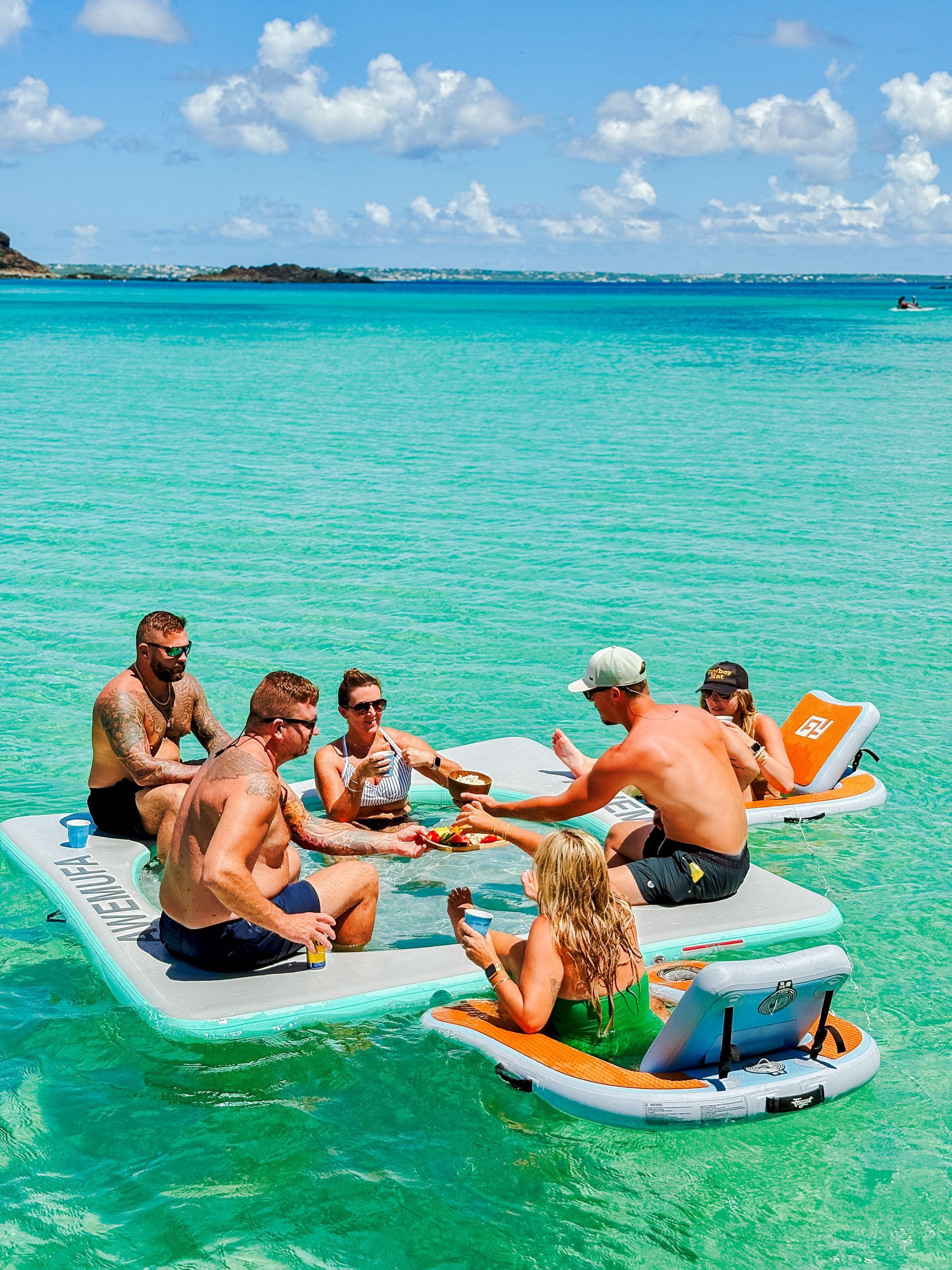 Group of people enjoying drinks on floating platforms in clear blue water.