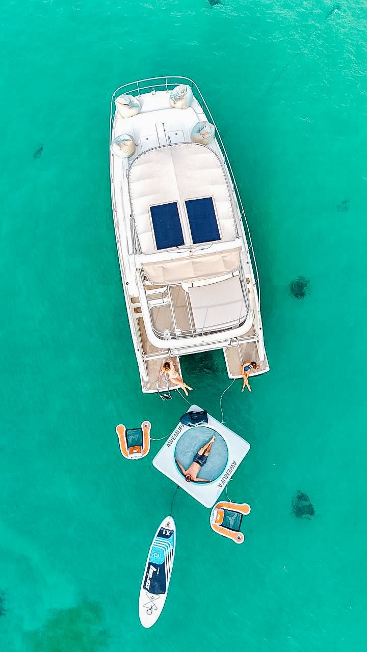 Aerial view of yacht and floating platform in turquoise water. People sunbathe, a paddleboard is nearby.