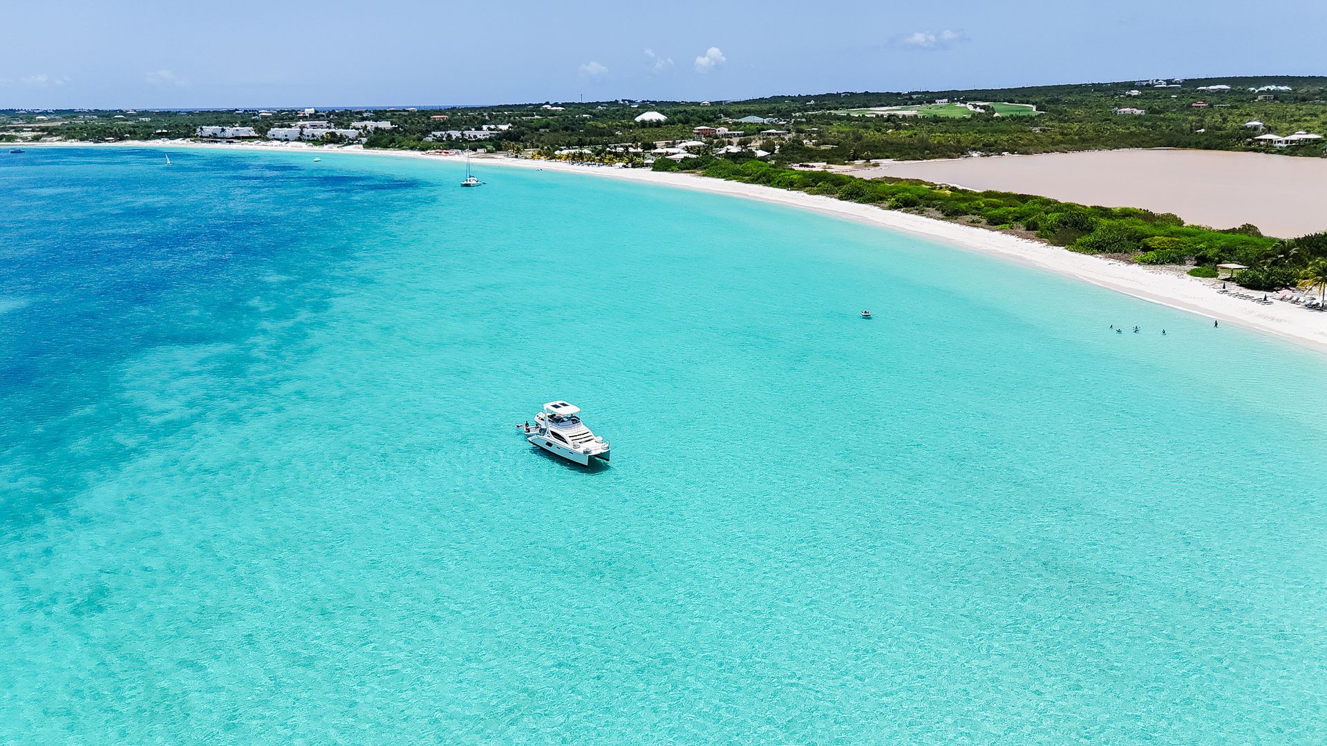 Turquoise ocean, white sand beach, boat, and lush green shoreline under a bright blue sky.