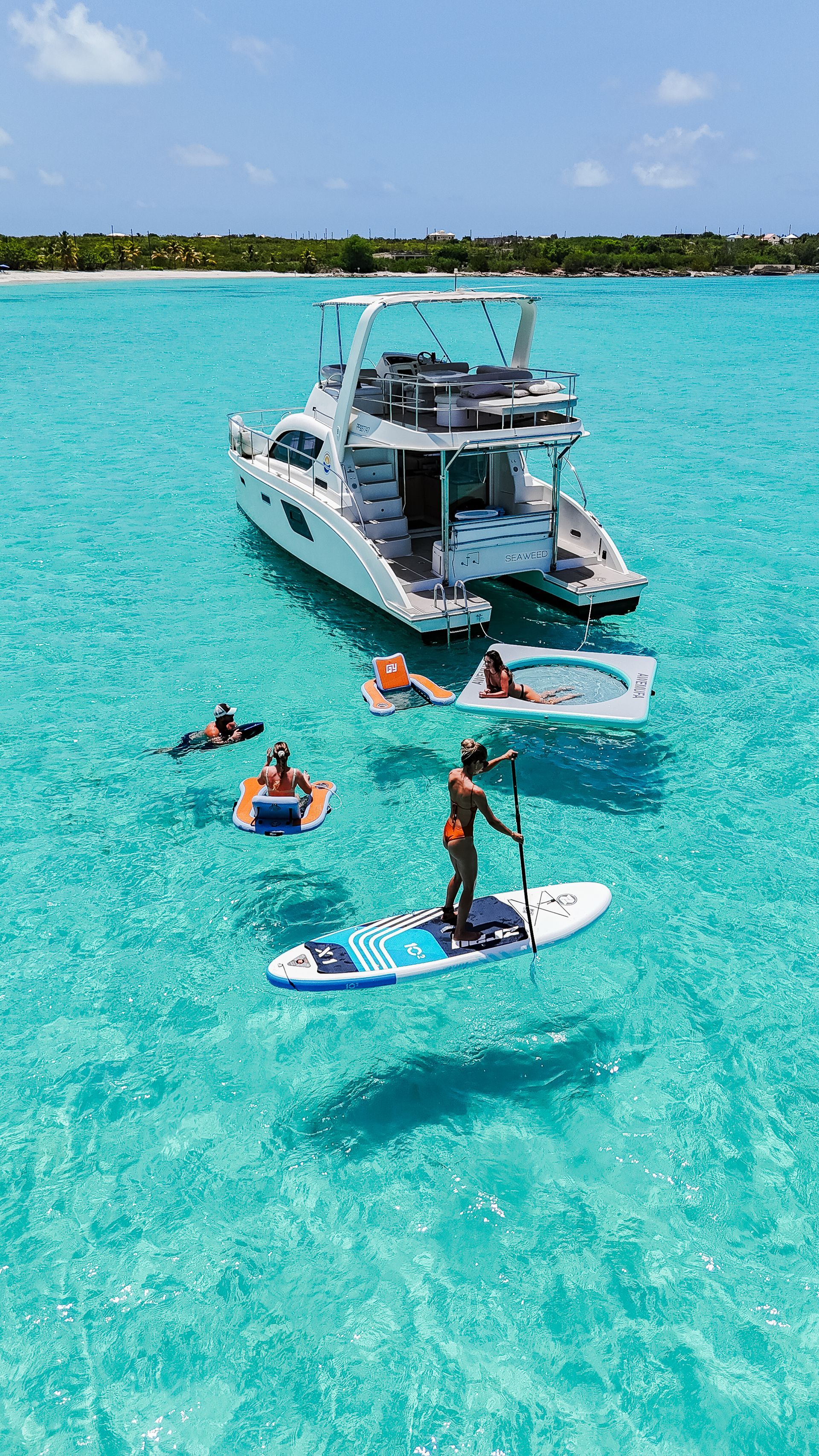 Catamaran in turquoise water with people swimming, paddleboarding, and floating. Sunny beach backdrop.