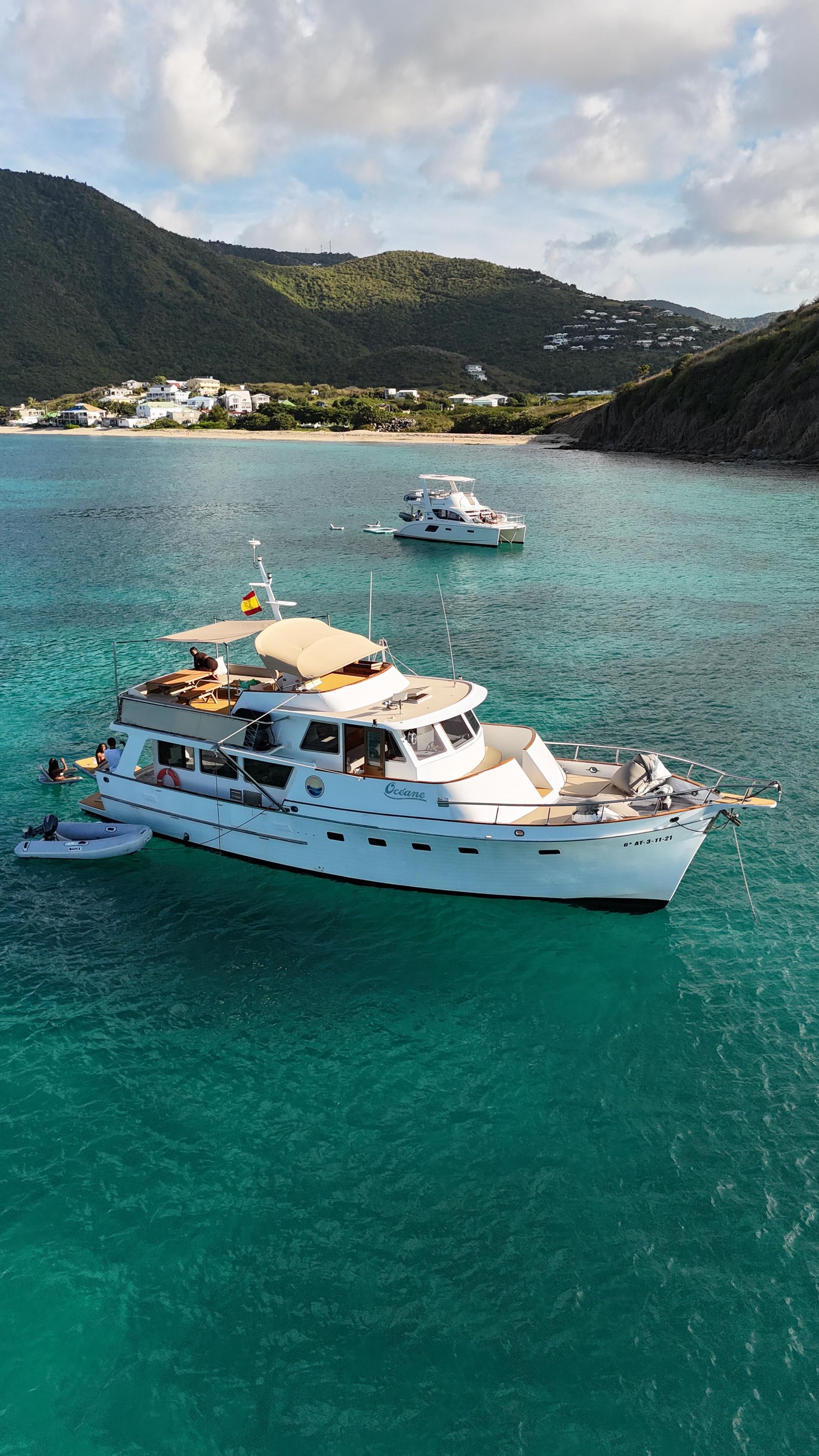 White yacht on turquoise water, with a smaller boat alongside, mountains in the background.