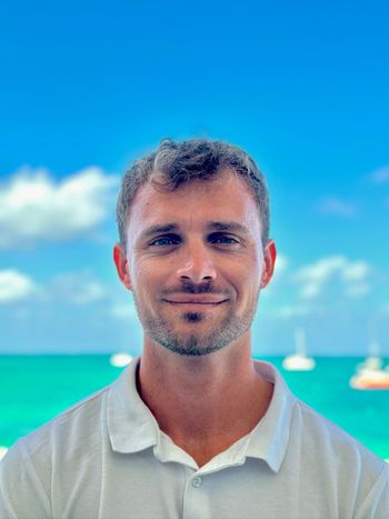 Man smiling in front of a turquoise ocean and blue sky with sailboats.
