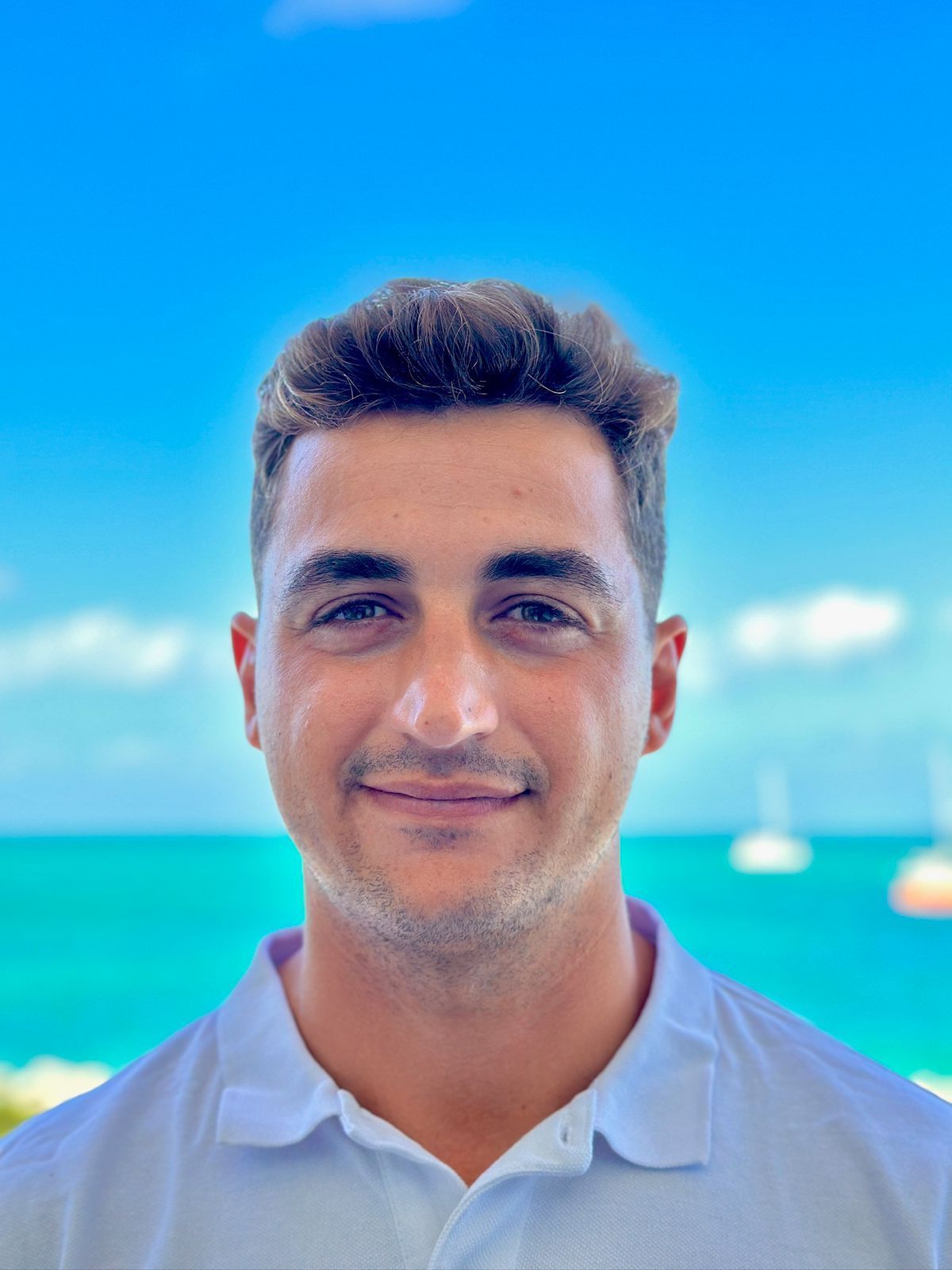 Man smiling, blue sky and ocean background. Wearing a white polo shirt, sunny outdoor setting.