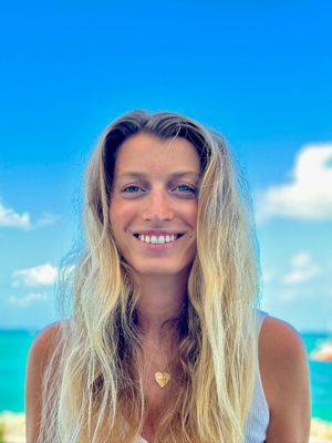 Woman with long blonde hair smiles in front of a blue sky and water. She wears a heart-shaped necklace.