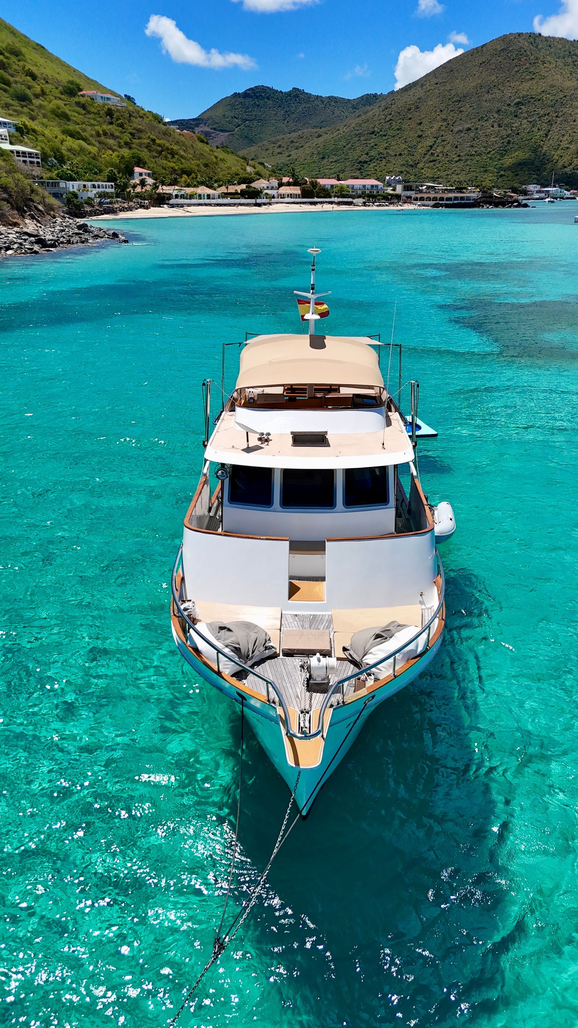 White yacht floats in turquoise water with a bright blue sky, near a beach and hillside.