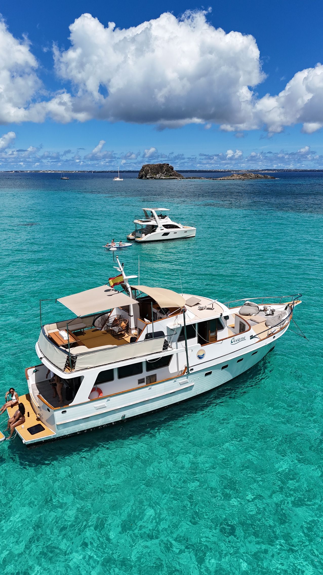 White yacht on turquoise water, with a smaller boat alongside, mountains in the background.