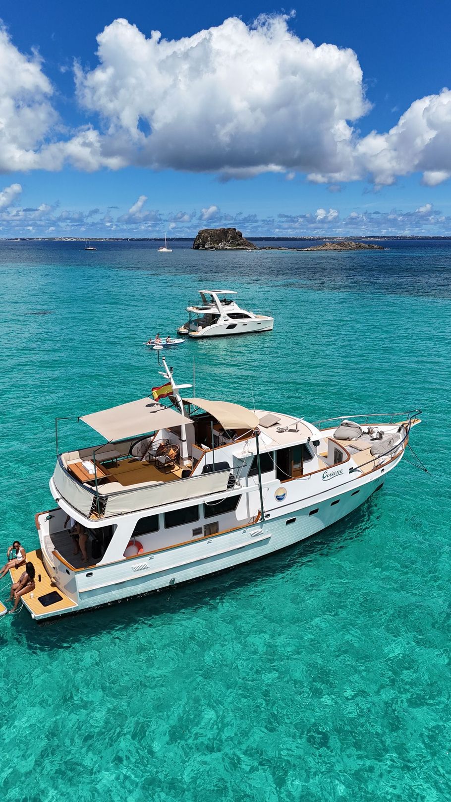 White yacht on turquoise water, with a smaller boat alongside, mountains in the background.