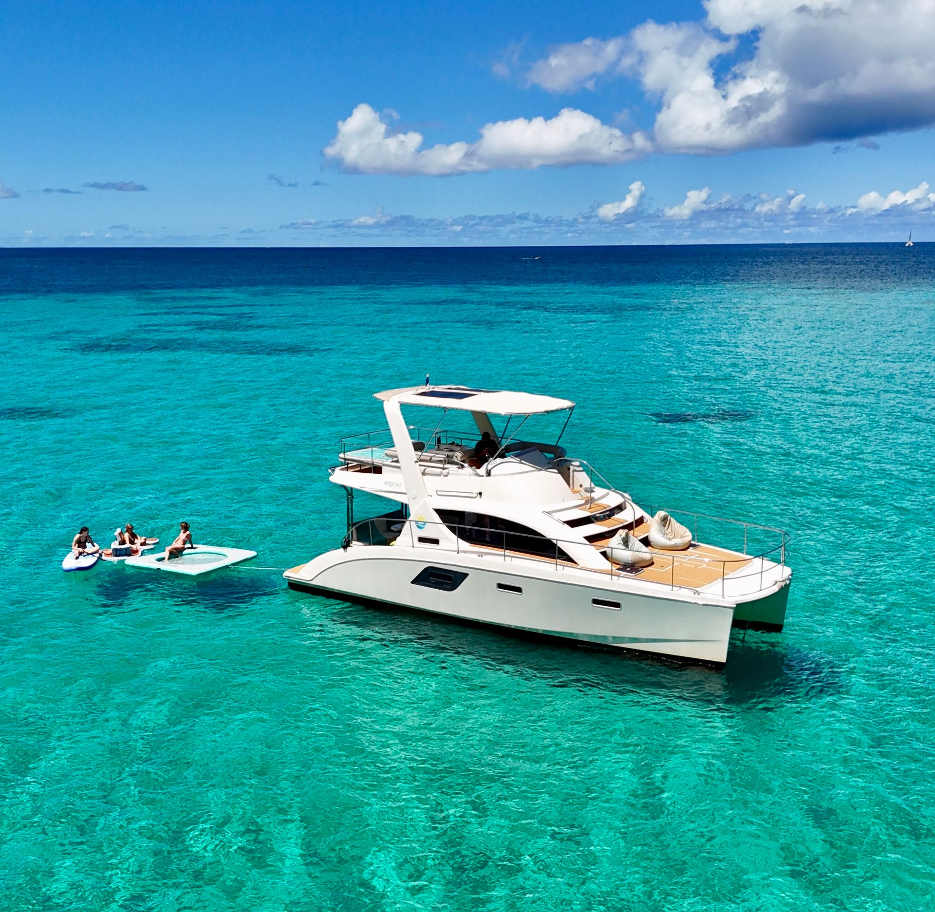White catamaran in turquoise water near a beach. People are near the boat with what appear to be paddleboards.