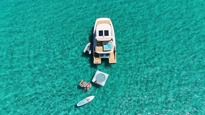 White catamaran in turquoise water with two people standing on the deck. Beach and green hills in background.