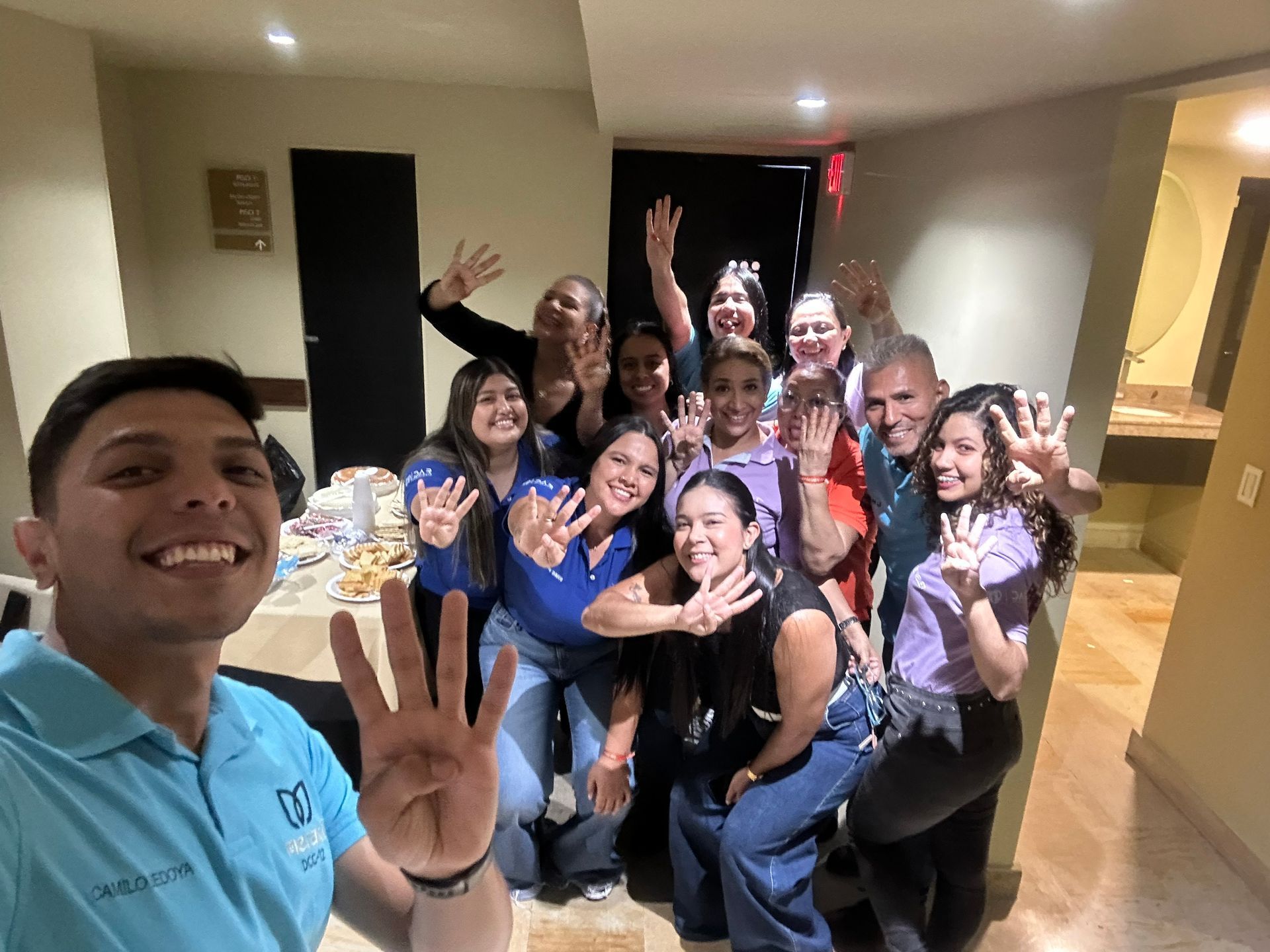 Un grupo de personas de pie en el interior de un edificio, sonriendo y levantando cuatro dedos