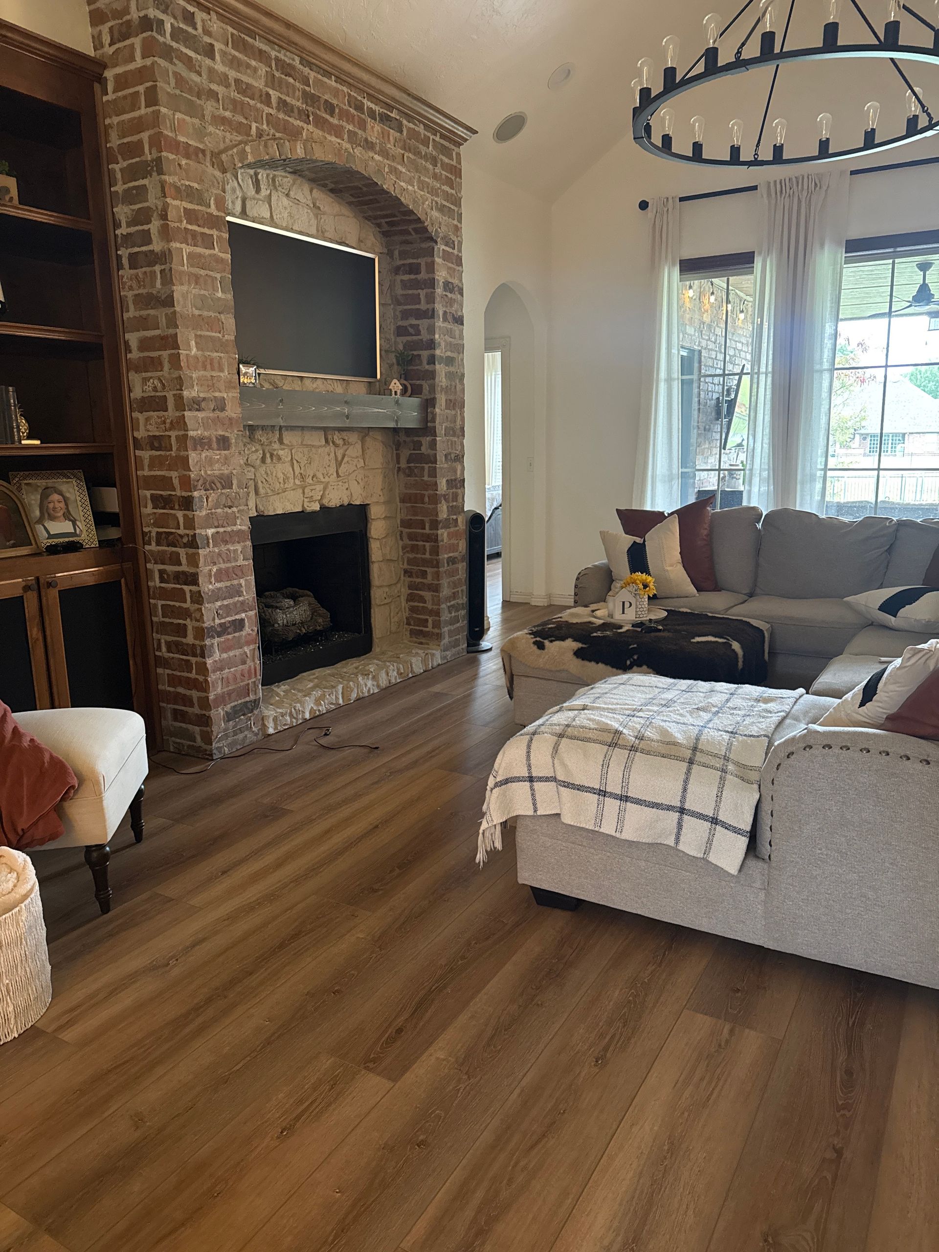 Bright living room with brick fireplace, large TV, sectional sofa, and a window with sheer curtains.