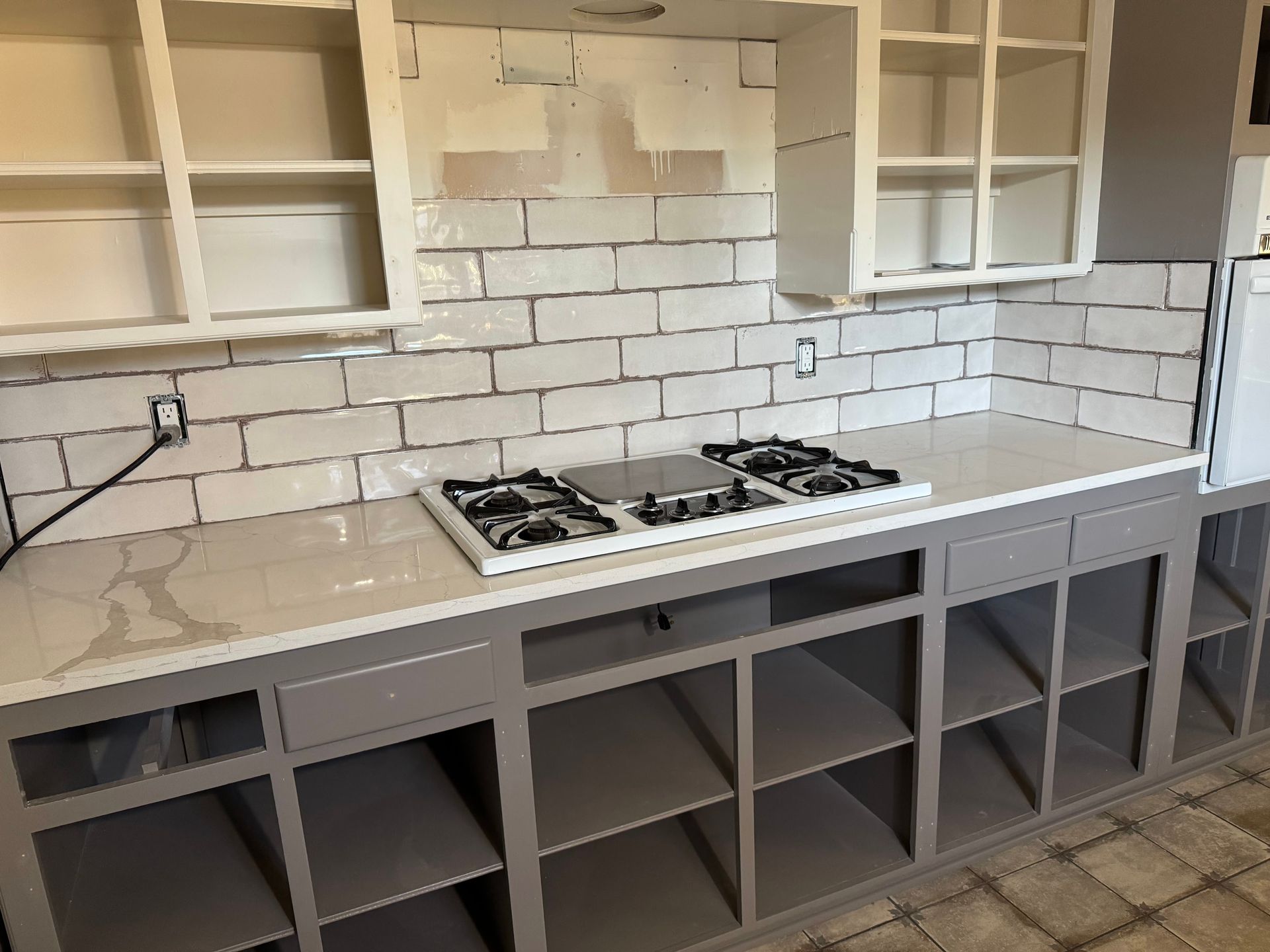 Modern kitchen with gray cabinets, white brick backsplash, and a gas cooktop under open shelves