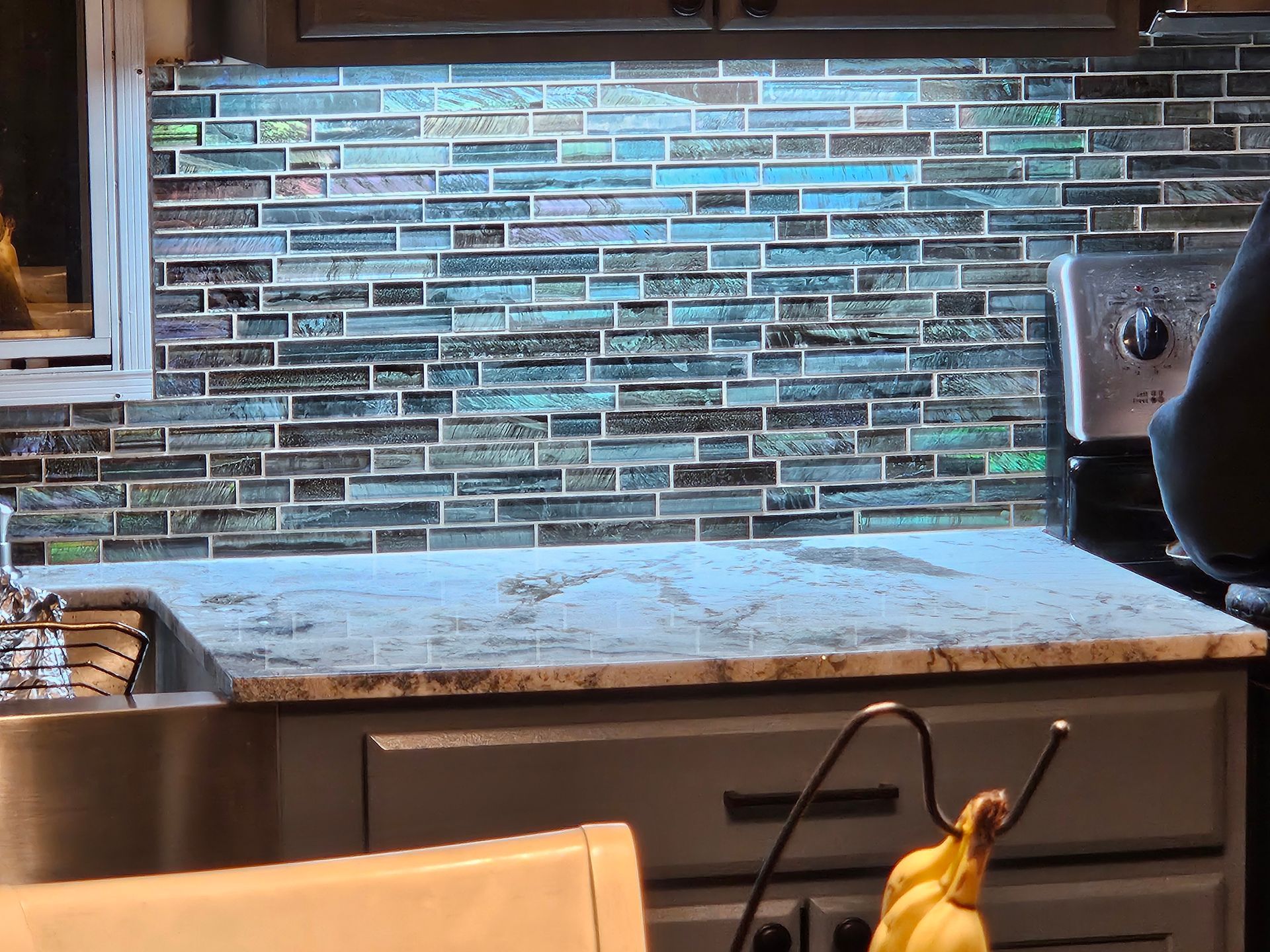 Kitchen backsplash with gray-blue tile, granite countertop, and stainless sink under cabinet lighting