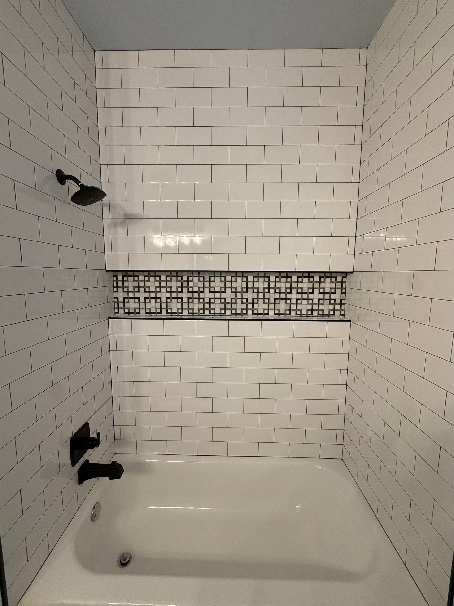 White-tiled bathroom shower with a bathtub, black fixtures, and decorative accent tile strip.