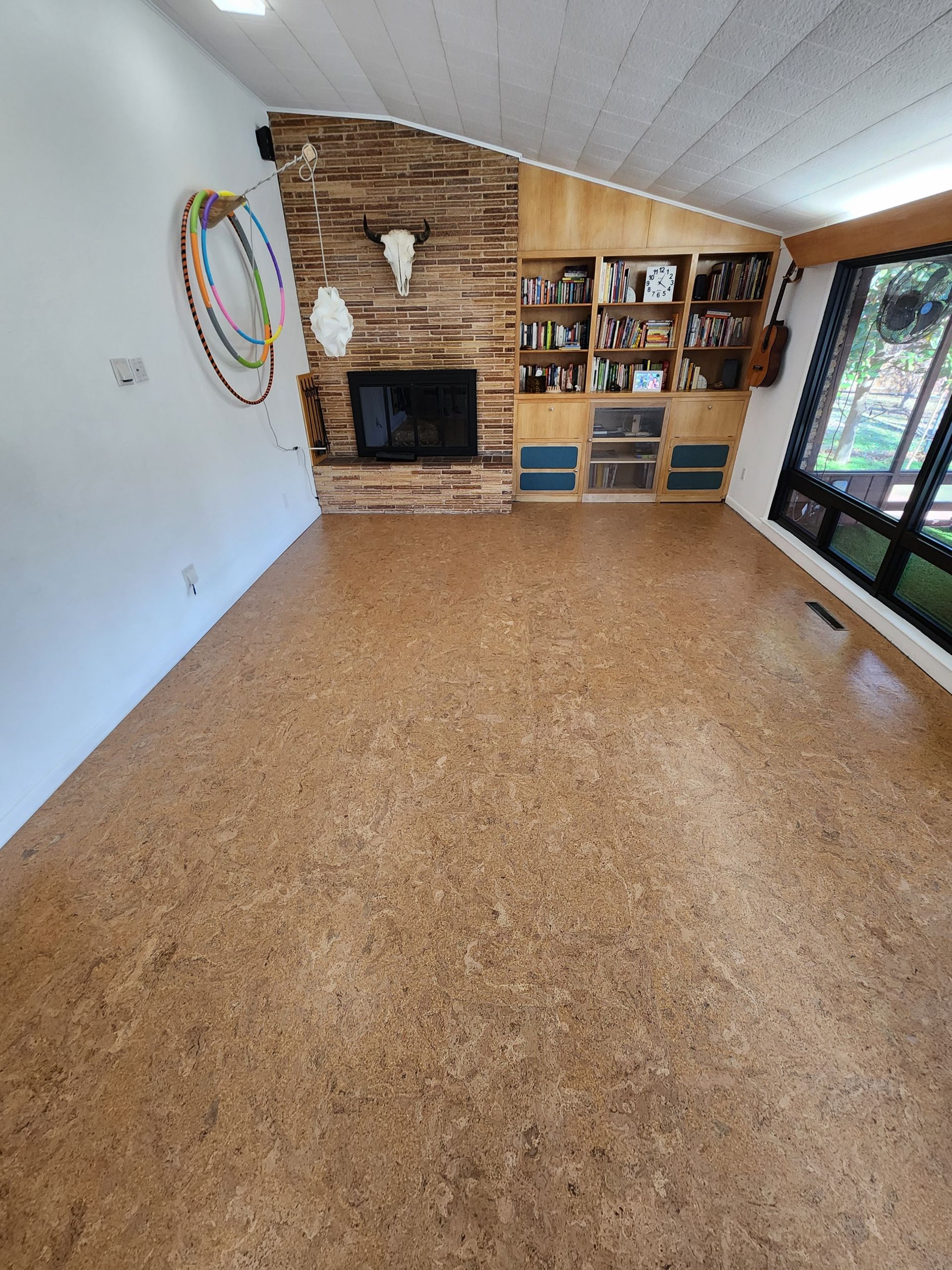 Empty living room with cork floor, stone fireplace, built-in shelves, and large windows.