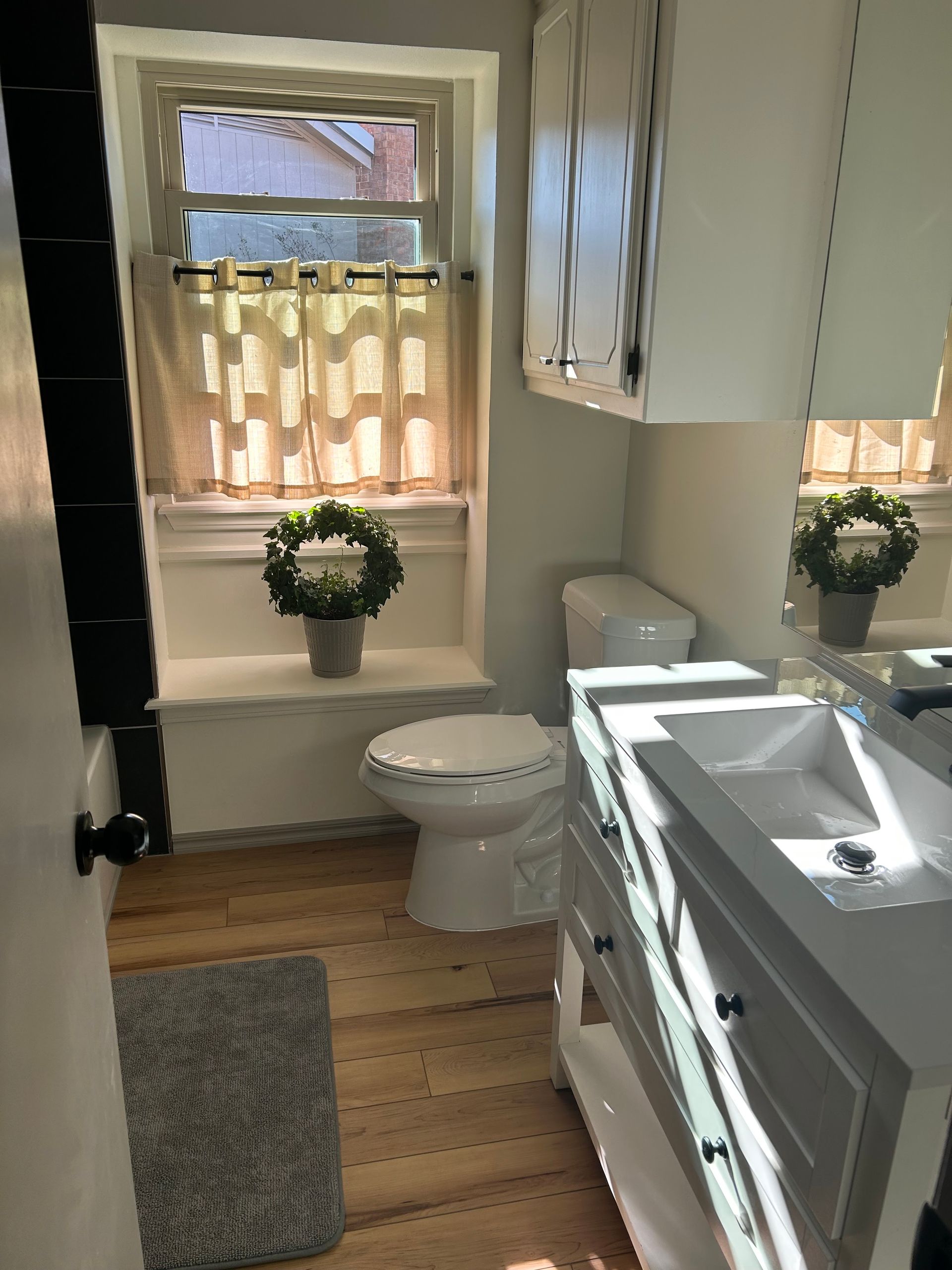 Small bathroom with white vanity, toilet, window, wood floor, and gray rug.