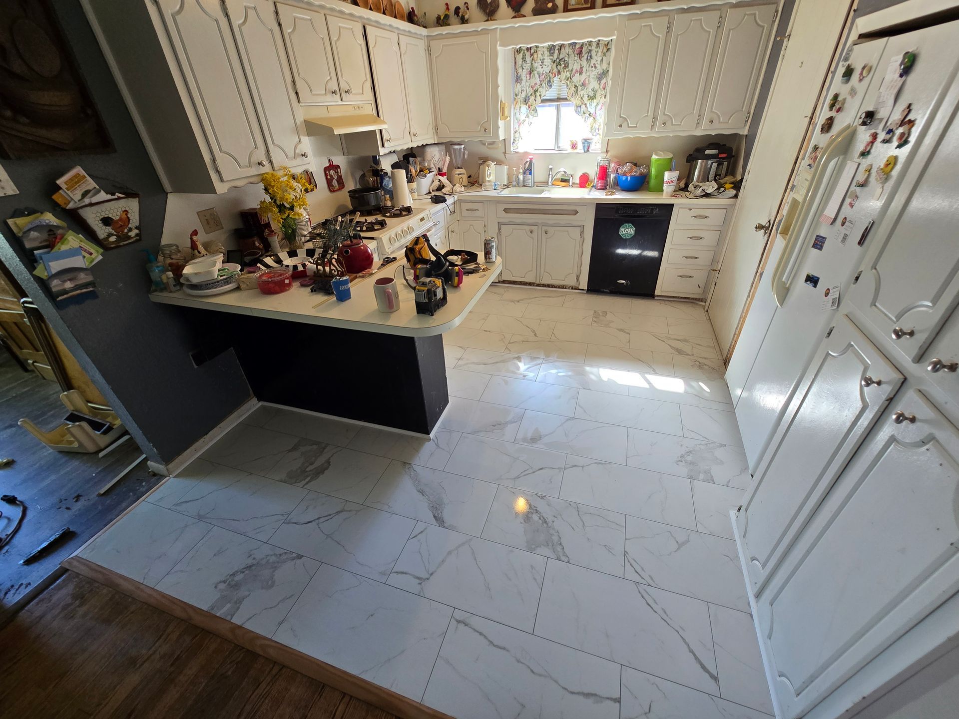 Bright kitchen with white cabinets, black countertops, tiled floor, and a central island cluttered with items