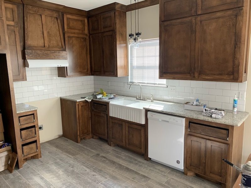 Empty kitchen with dark wood cabinets, white tile backsplash, sink under window, and gray wood flooring