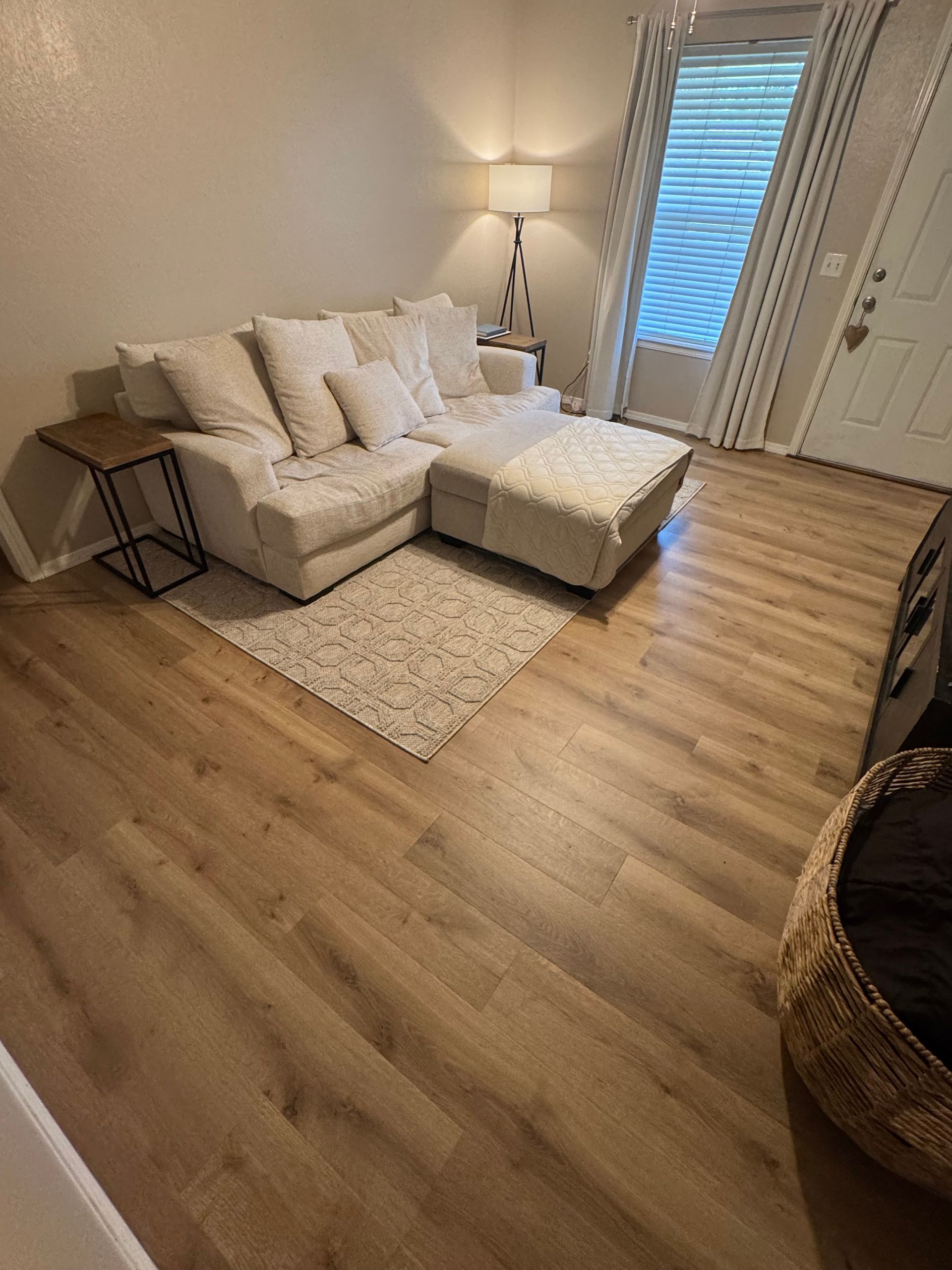 Cozy beige living room with sectional sofa, rug, lamp, and hardwood floor beside a window with blinds