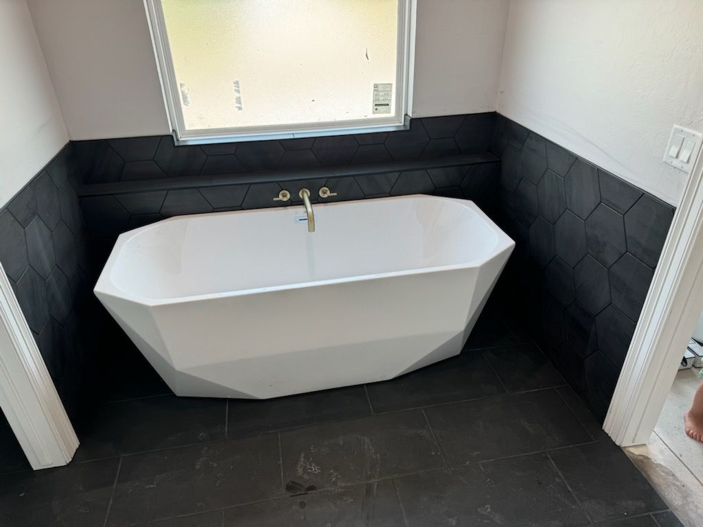 Modern freestanding white bathtub in a black-tiled bathroom beneath a window