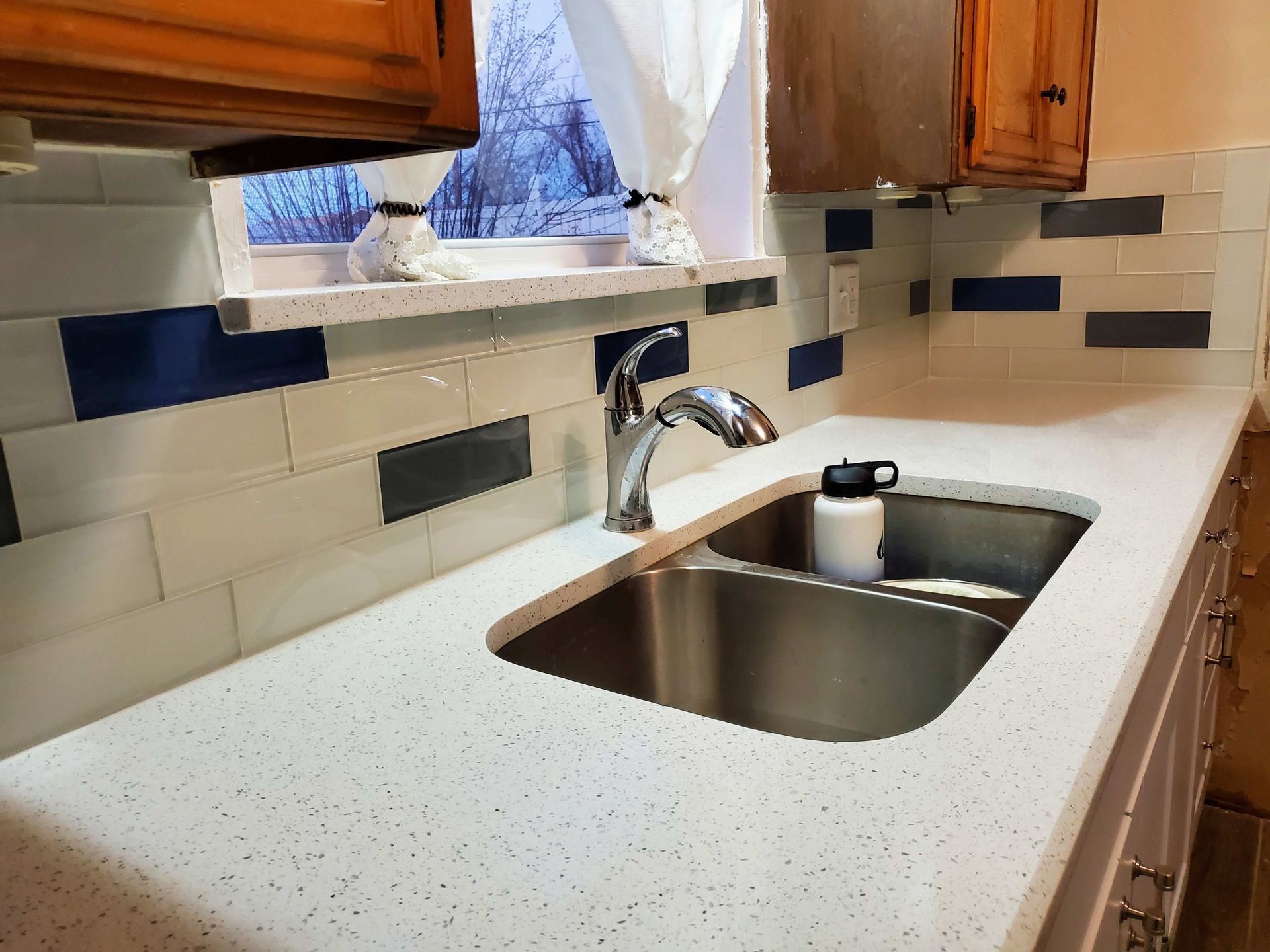 Kitchen countertop with double sink, chrome faucet, and tiled backsplash under wooden cabinets