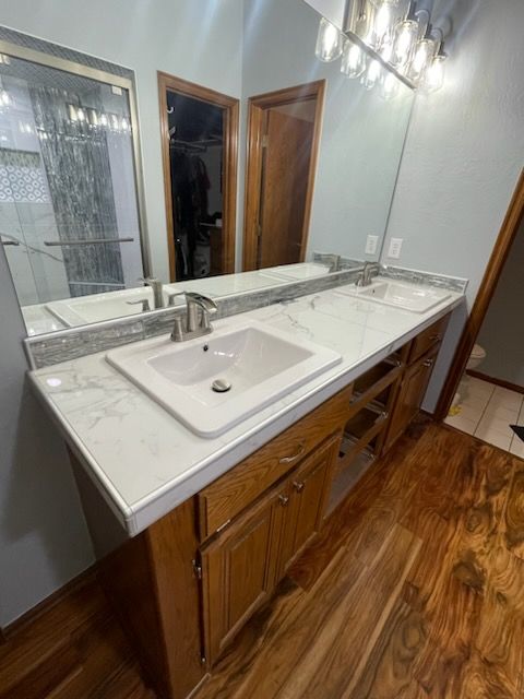 Bathroom vanity with white countertop, dual sinks, mirror, and wooden cabinets beside a wood floor