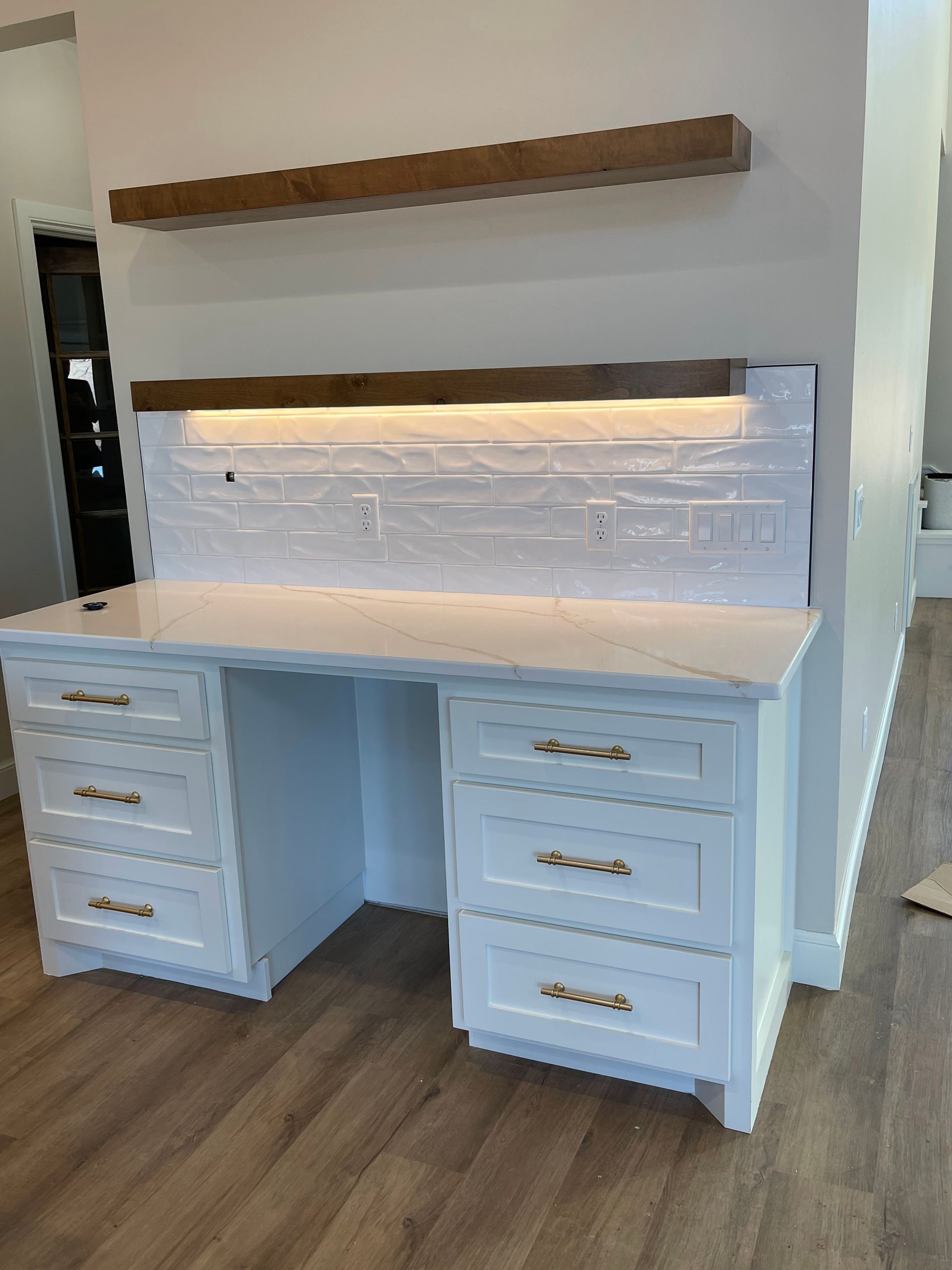 White built-in desk with drawers, wood shelf above, and LED-lit backsplash in a modern room