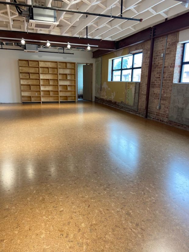 Empty studio with polished floor, exposed brick walls, large windows, and wall-mounted shelves.