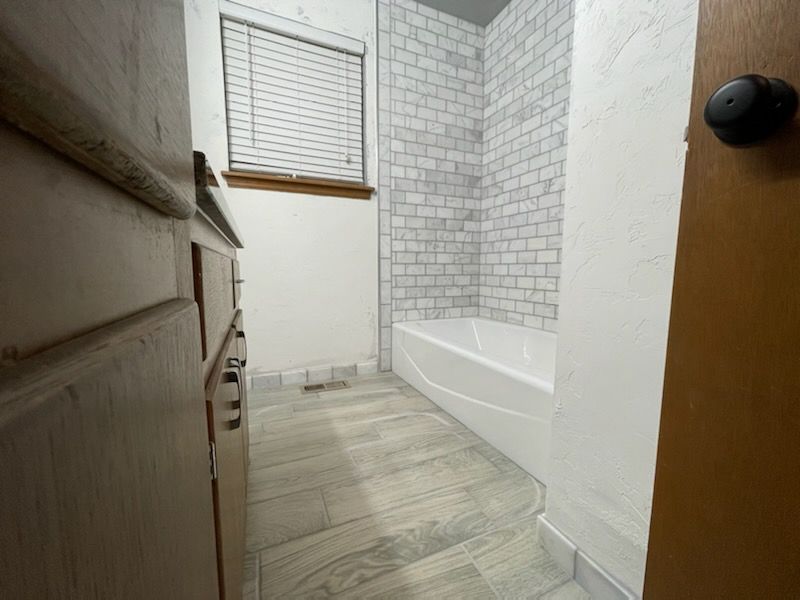 Narrow bathroom with white tile, bathtub, window, and wood-look floor.