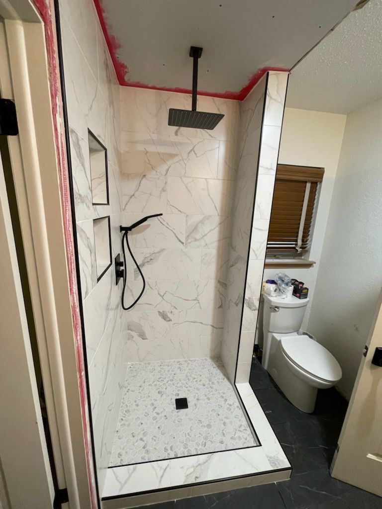 Partially finished tiled shower stall with black showerhead and white marble-look walls, beside a toilet.