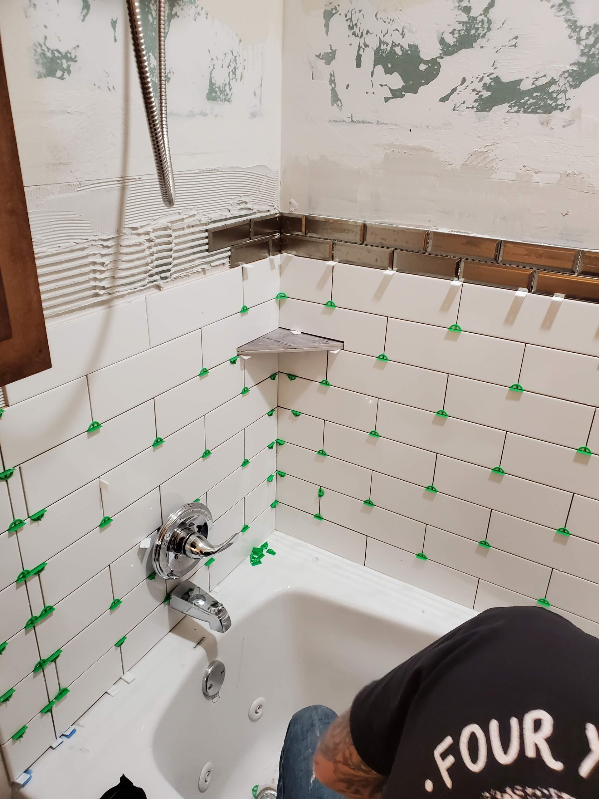 Bathroom under renovation with tiled shower wall, exposed plumbing, and a worker crouching in the tub.