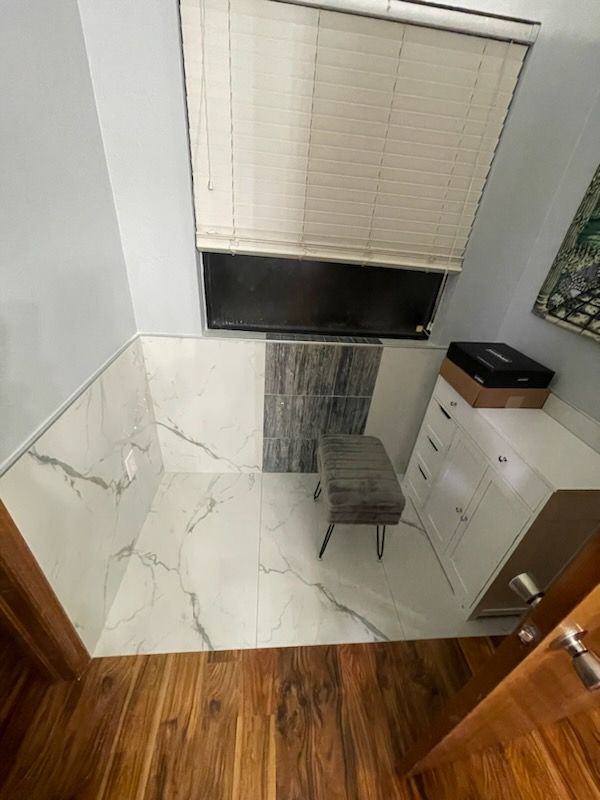 Small tiled room with a window, blinds, stool, and white cabinets beside a wooden door.