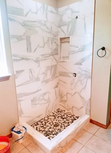 Bathroom shower with marble-pattern tile walls, white curb, and pebble floor accent.