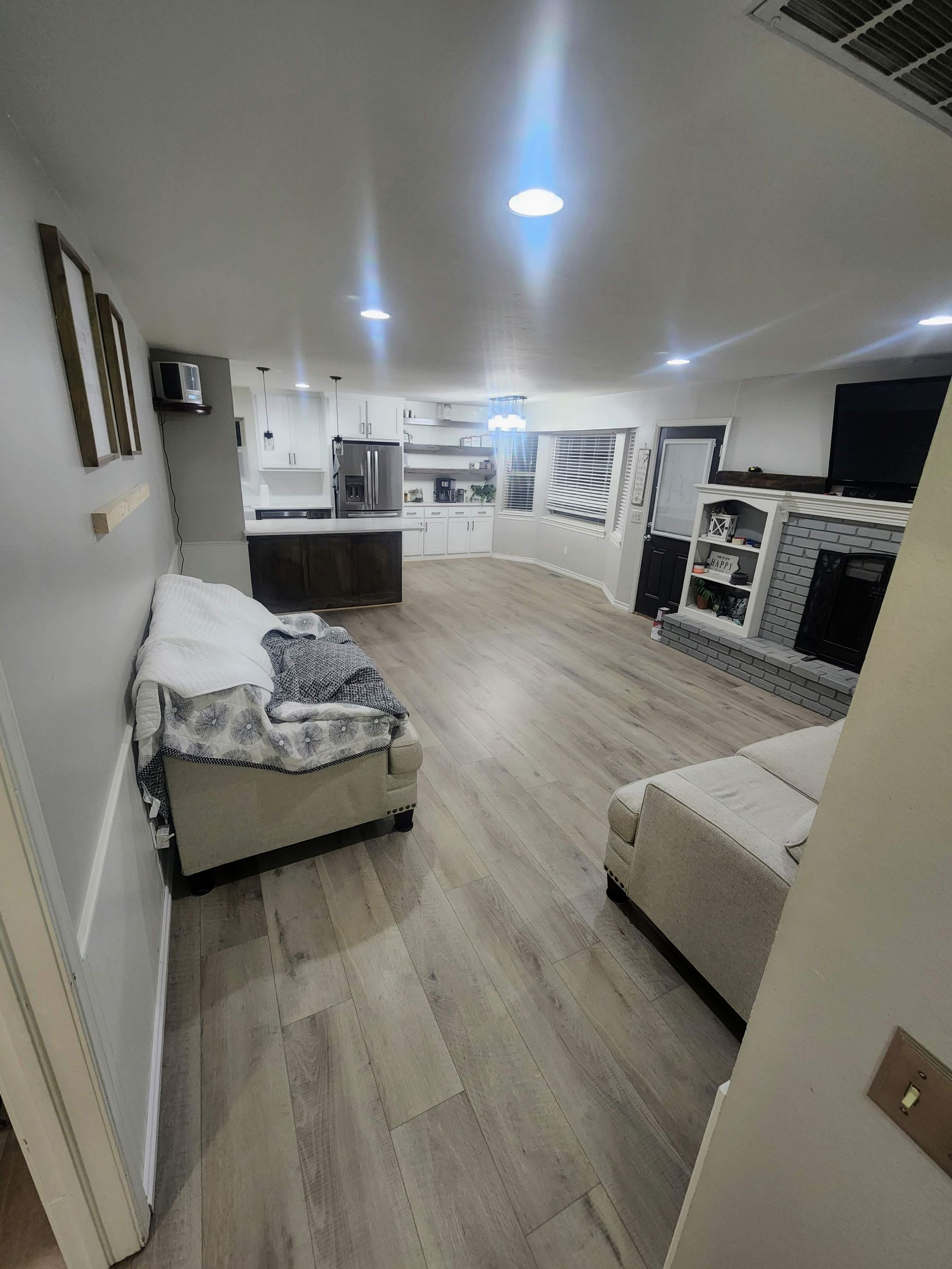 Open-concept living room and kitchen with light wood floors, beige furniture, and bright ceiling lights