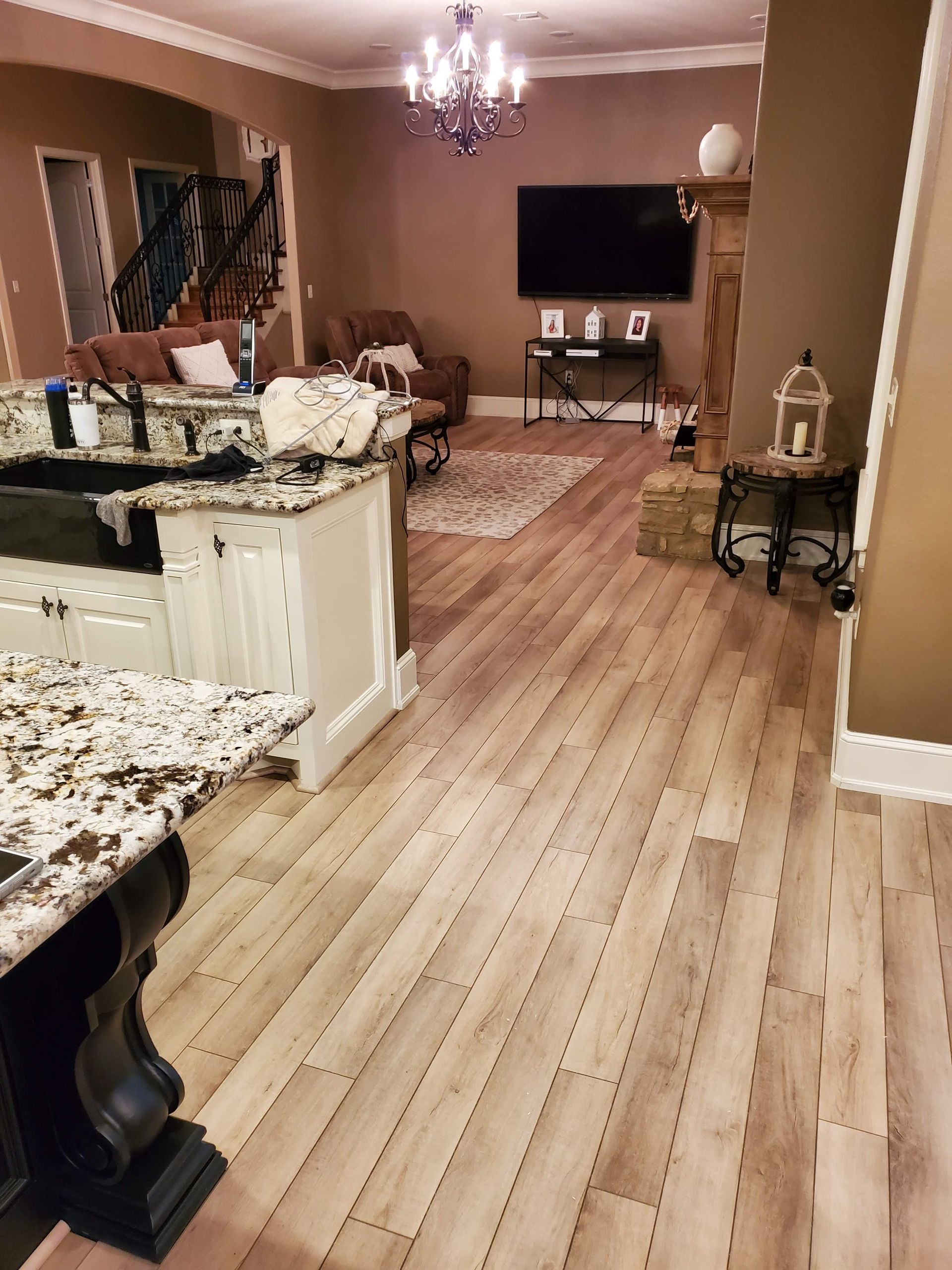 Open-plan kitchen and living room with wood floors, granite counters, chandelier, wall-mounted TV, and patterned rug.