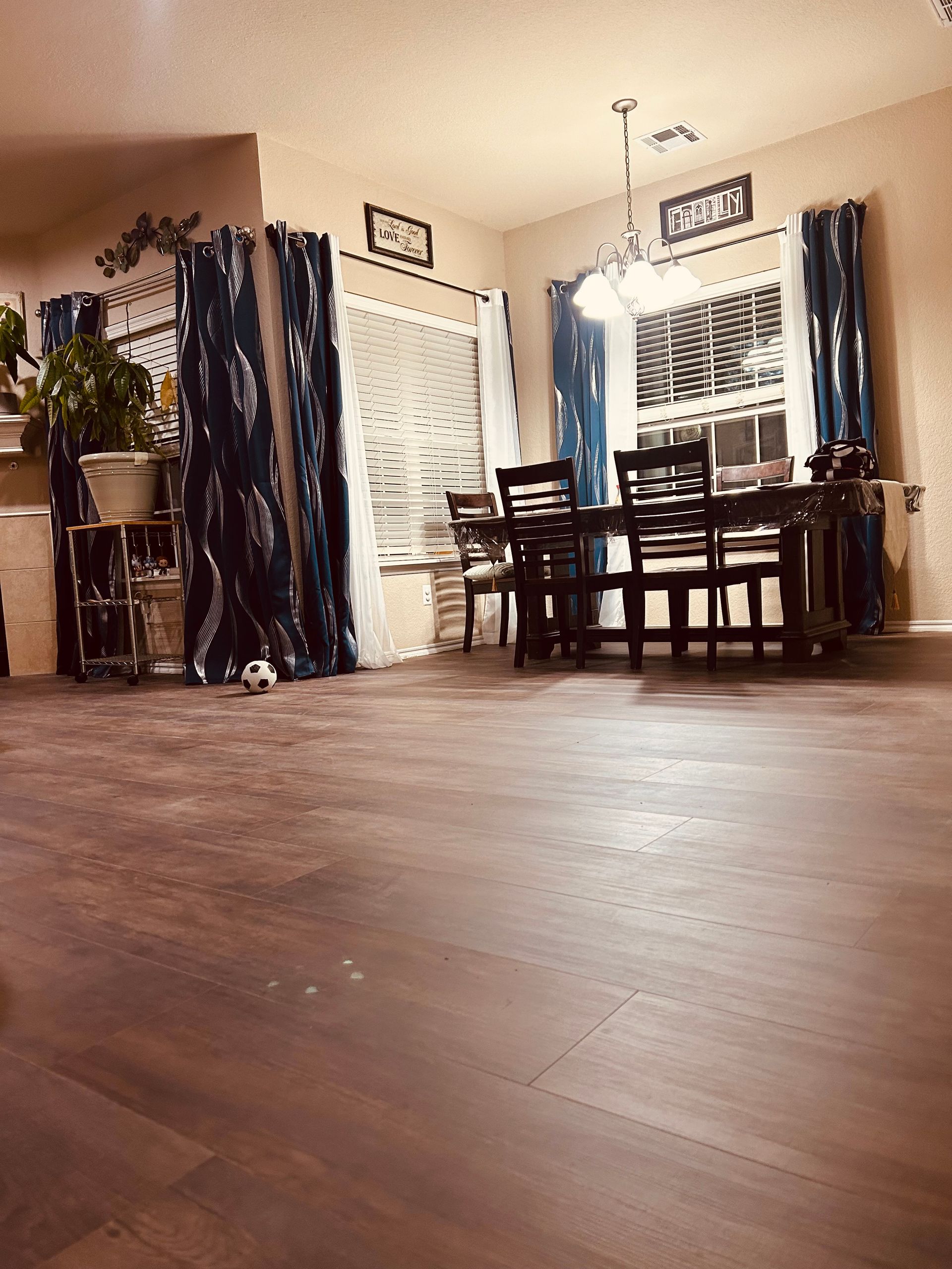 Dining room with dark wood floor, table and chairs, windows with curtains, and potted plants in the corners