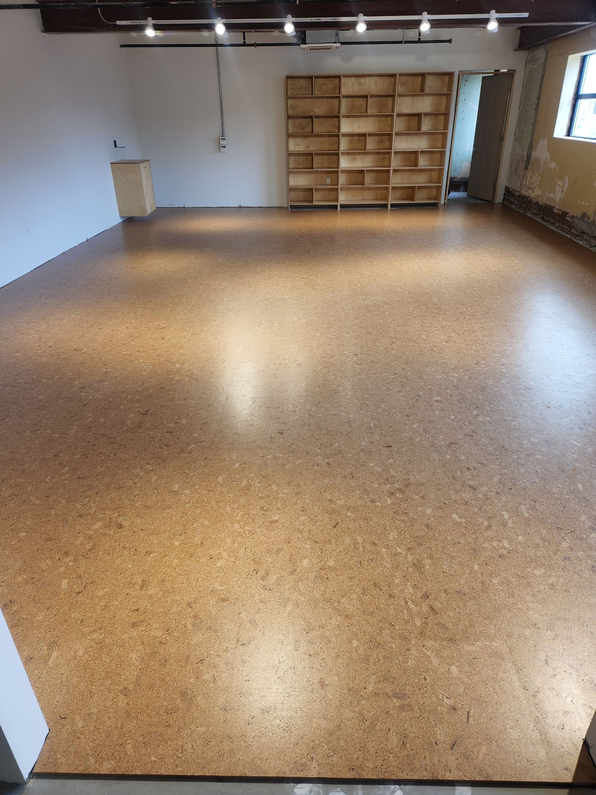 Empty room with speckled floor, overhead lights, wooden shelves, and a window on the right.