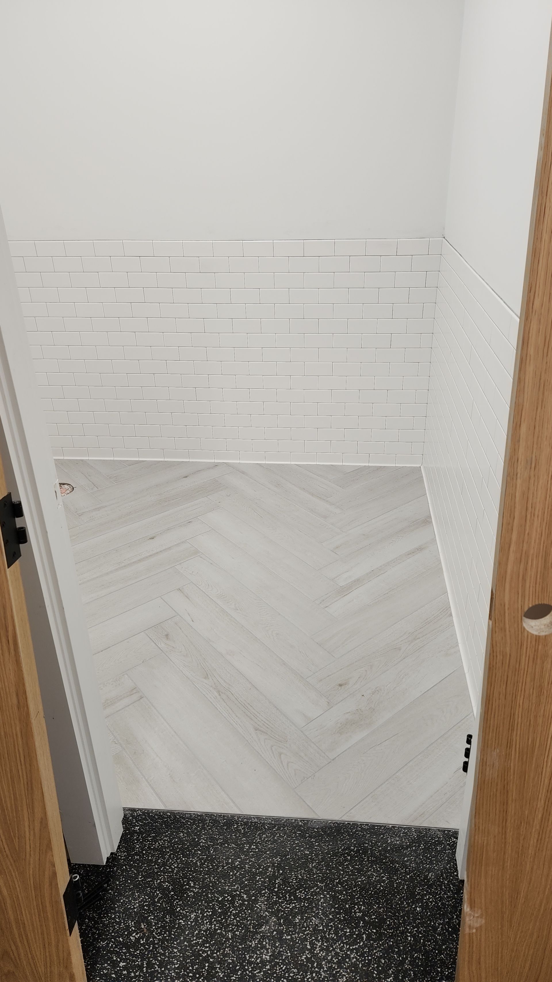 White tiled wall with a herringbone-patterned tile panel above a black-speckled floor, viewed through a doorway