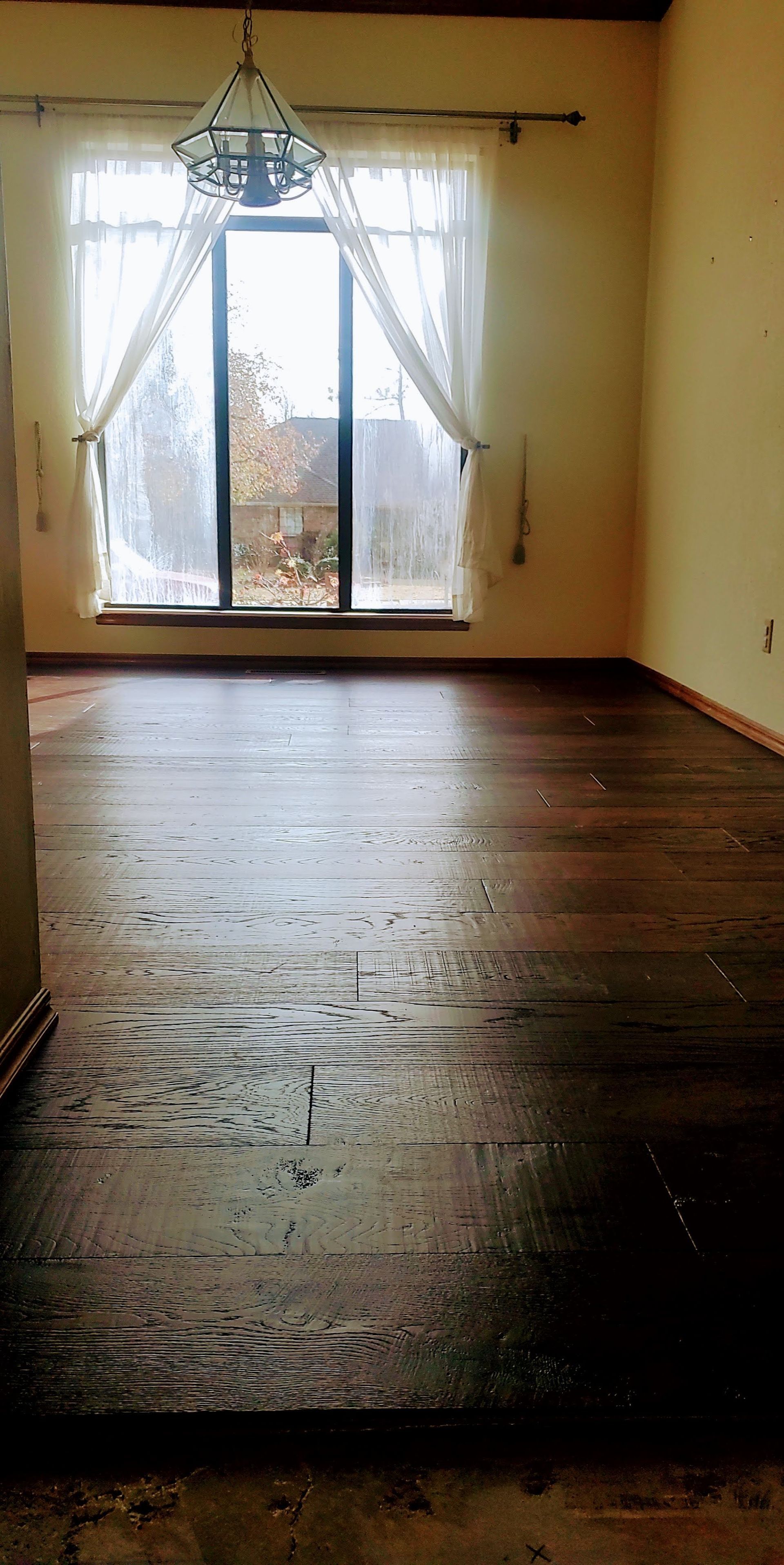Empty room with hardwood floor, chandelier, and sunlight through sheer curtains at the window