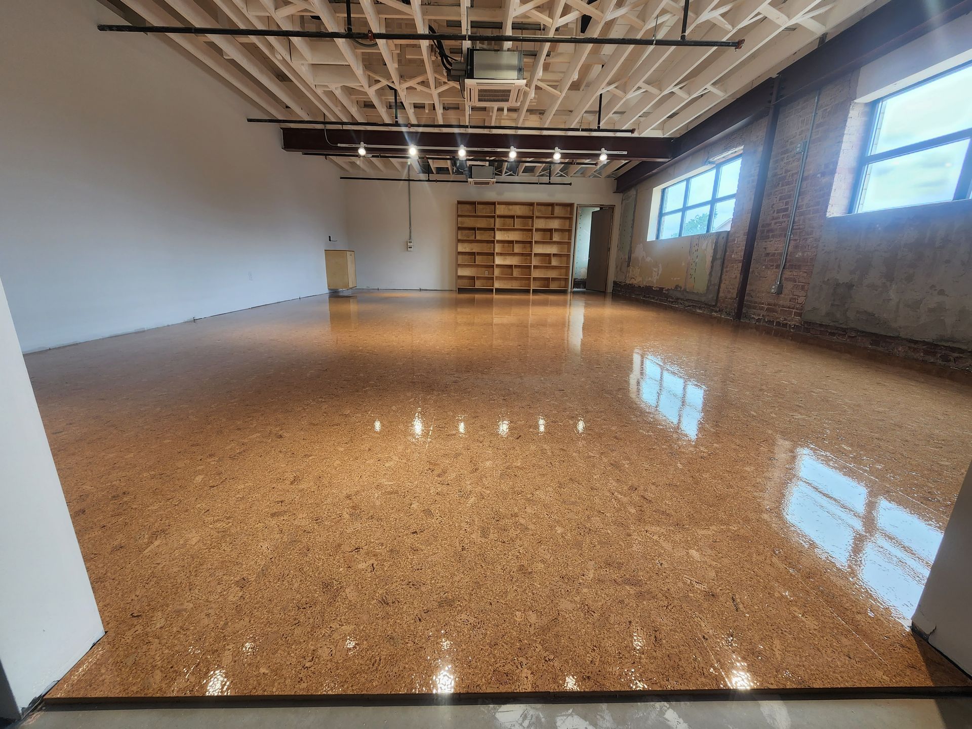 Empty polished workshop with cork floor, exposed ceiling beams, brick wall, and large windows.