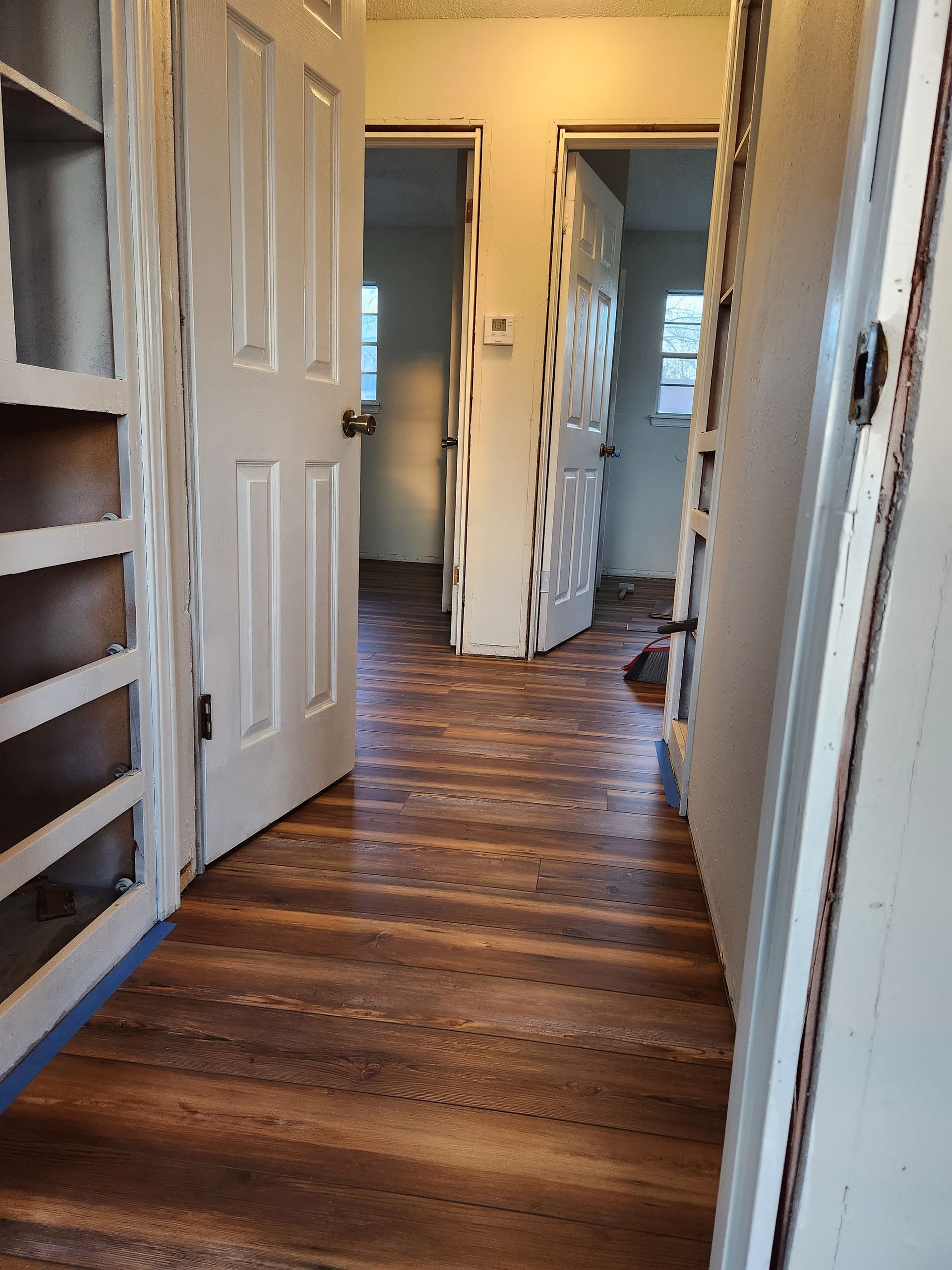 Narrow hallway with white doors, hardwood floors, and warm light from a room ahead.