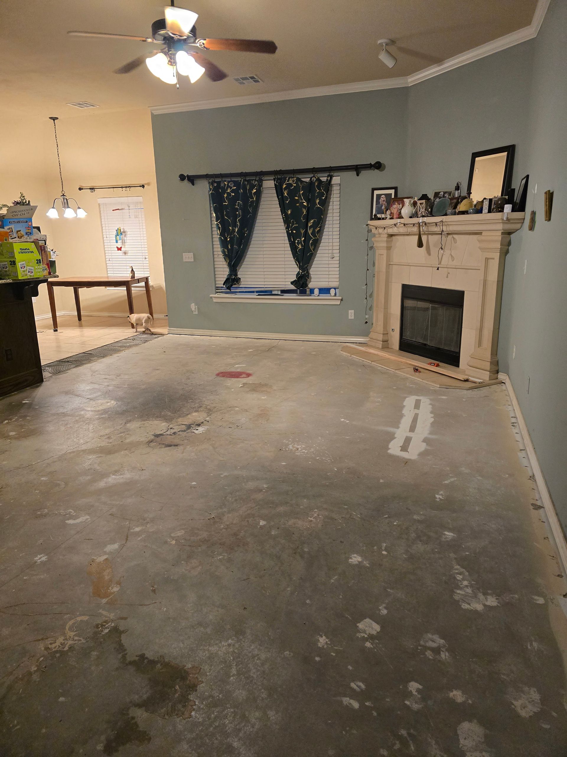 Empty living room with worn concrete floor, fireplace, window with dark curtains, and ceiling fan.