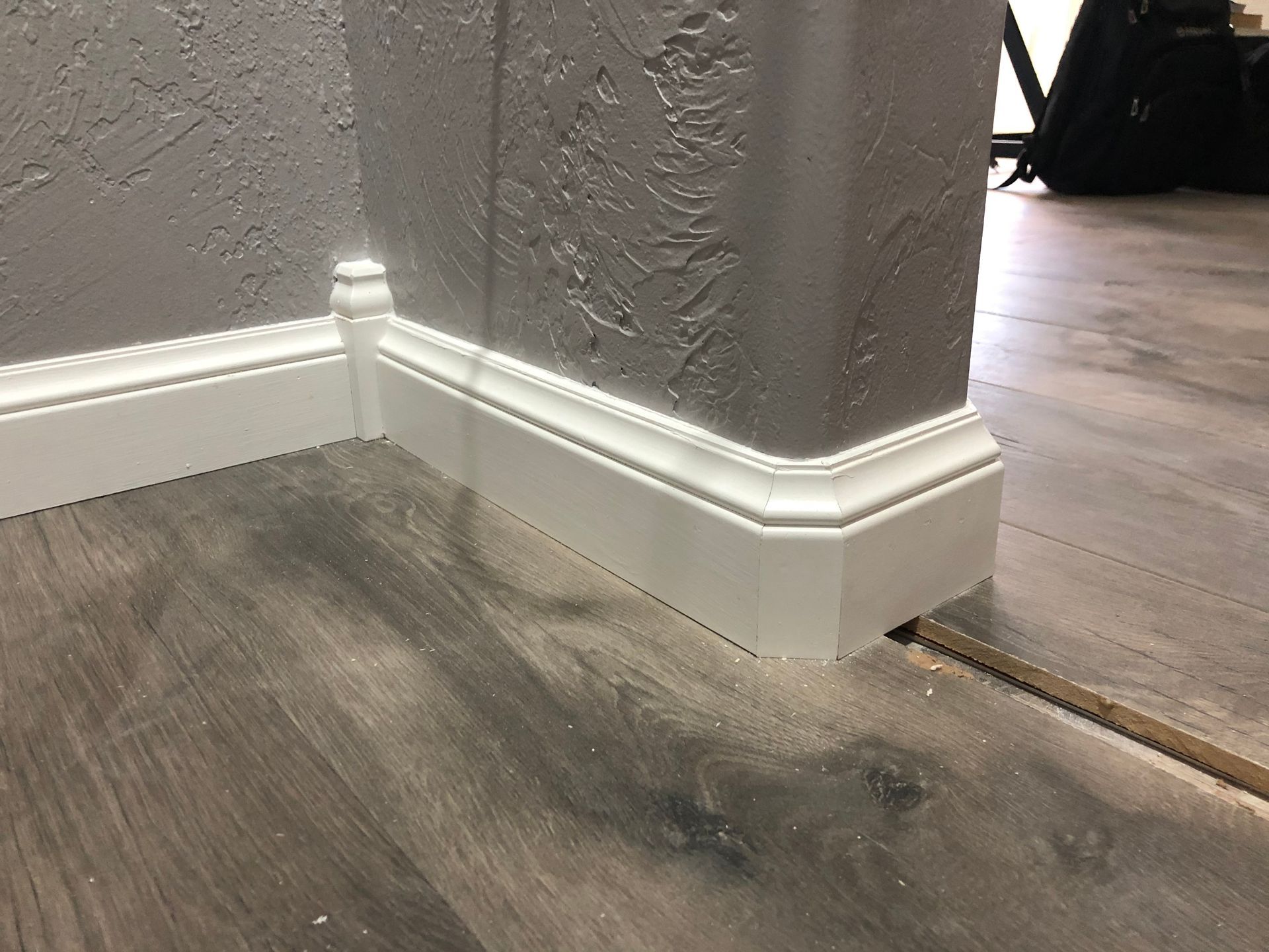 Baseboard corner where gray textured wall meets wood flooring and gray carpet with white trim.