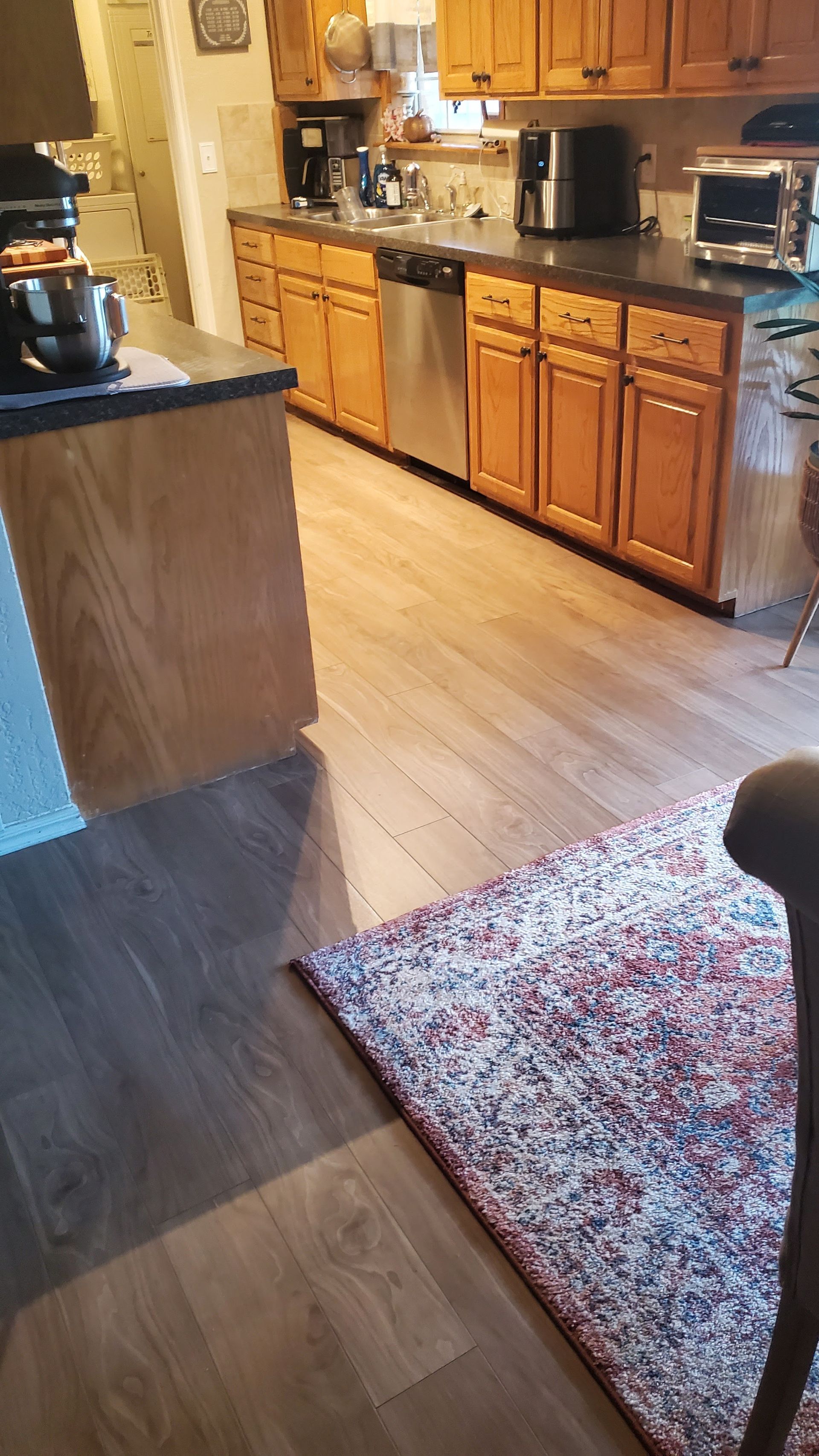 Kitchen with wooden cabinets, black countertop, and a patterned rug on a light wood floor.